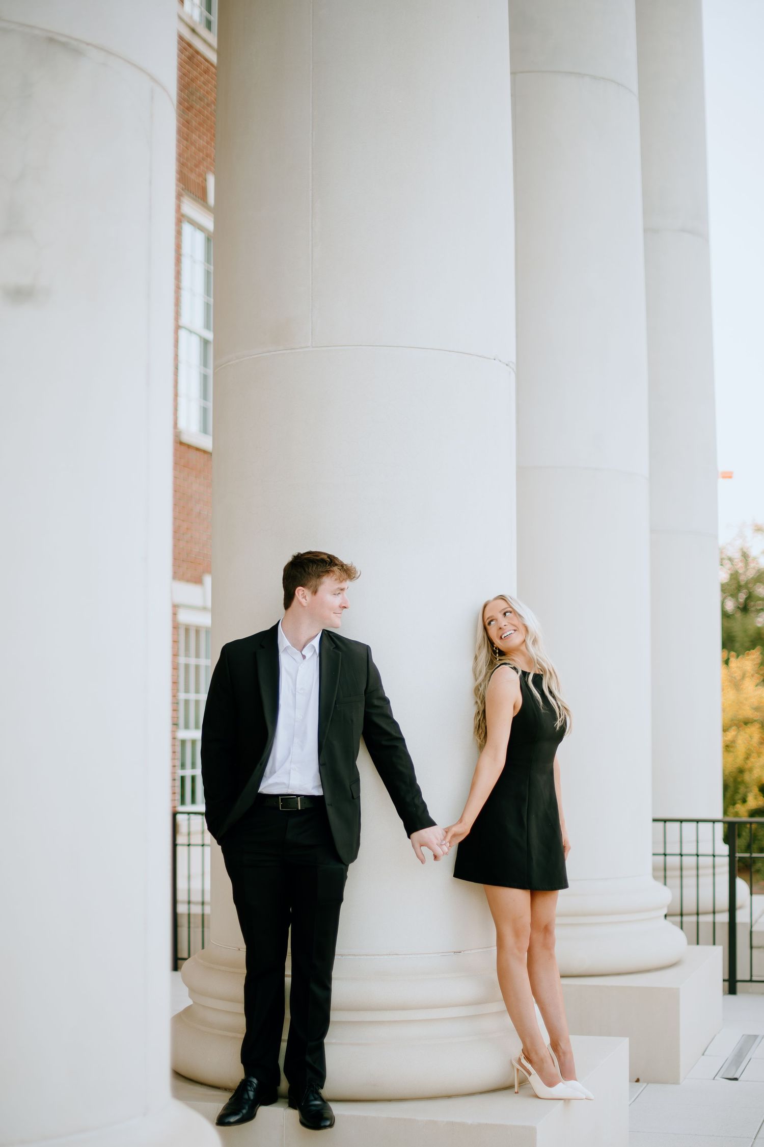 A couple in formal attire poses playfully between white columns on a porch.