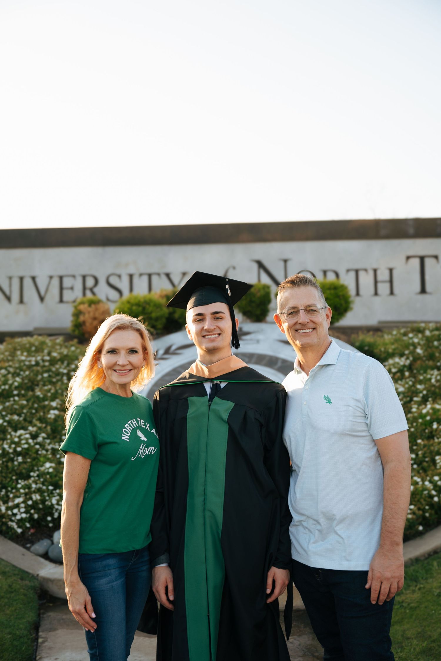 A graduate in green cap and gown poses with two proud family members in front of University of North Texas sign.