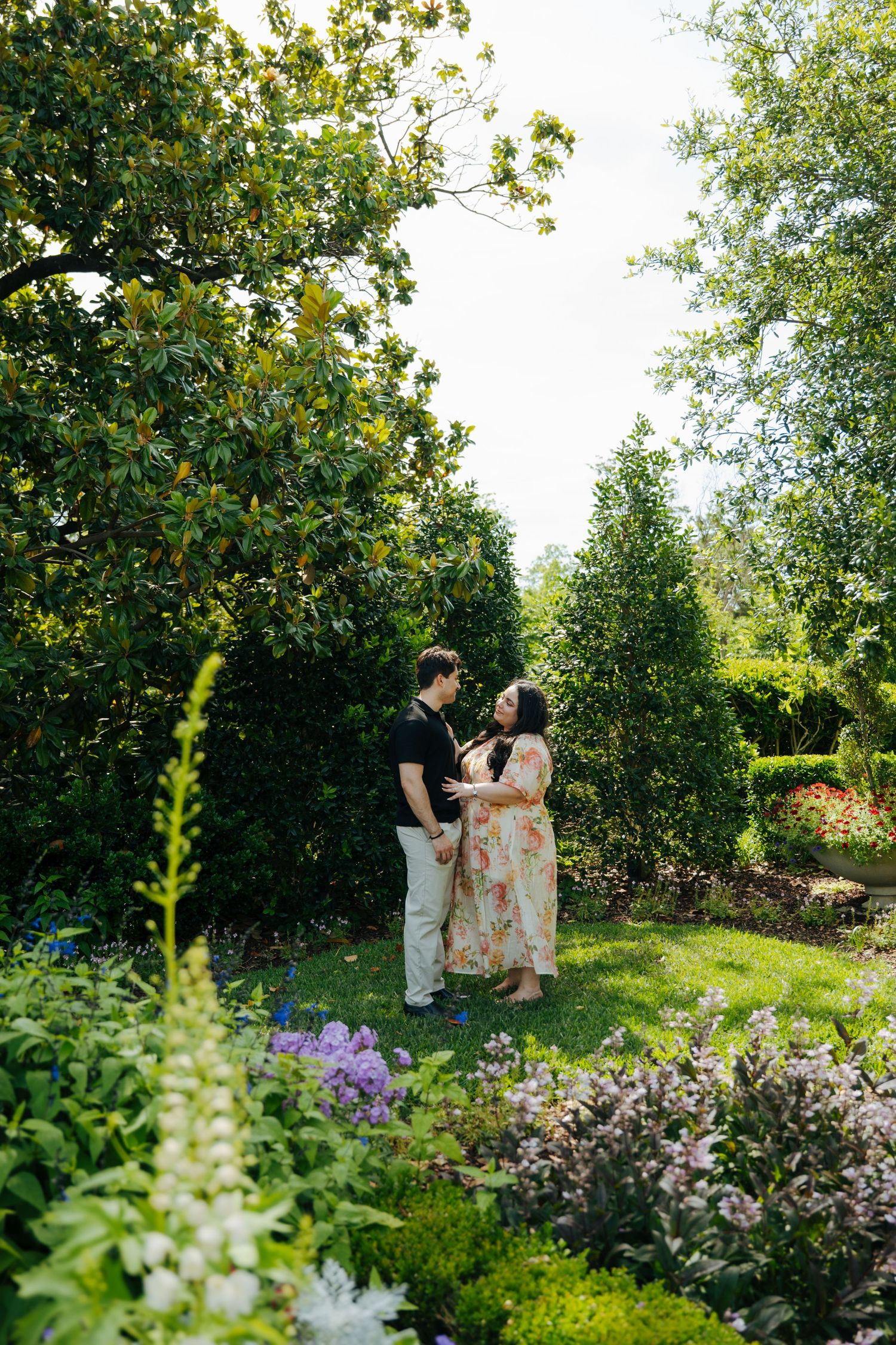 Couple holding hands and smiling in a lush garden surrounded by blooming flowers and tall green trees on a bright day.