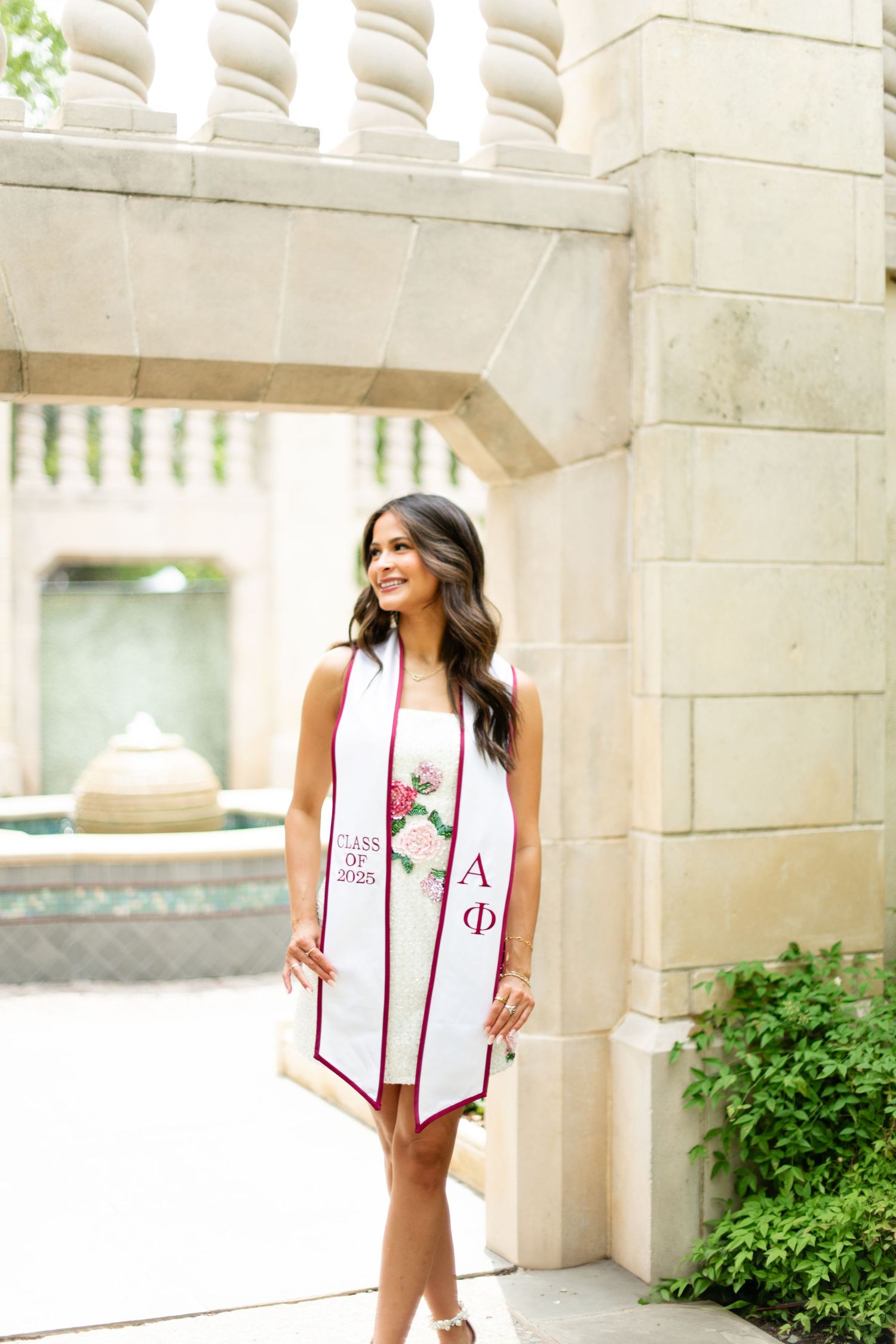 Graduate in white dress with decorative stole poses against classical stone architecture.