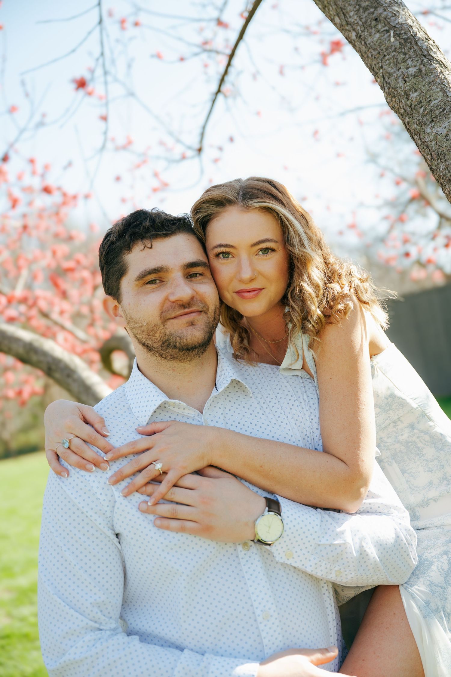 A couple embraces under a blooming pink tree, the woman hugging the man from behind as they smile warmly outdoors.