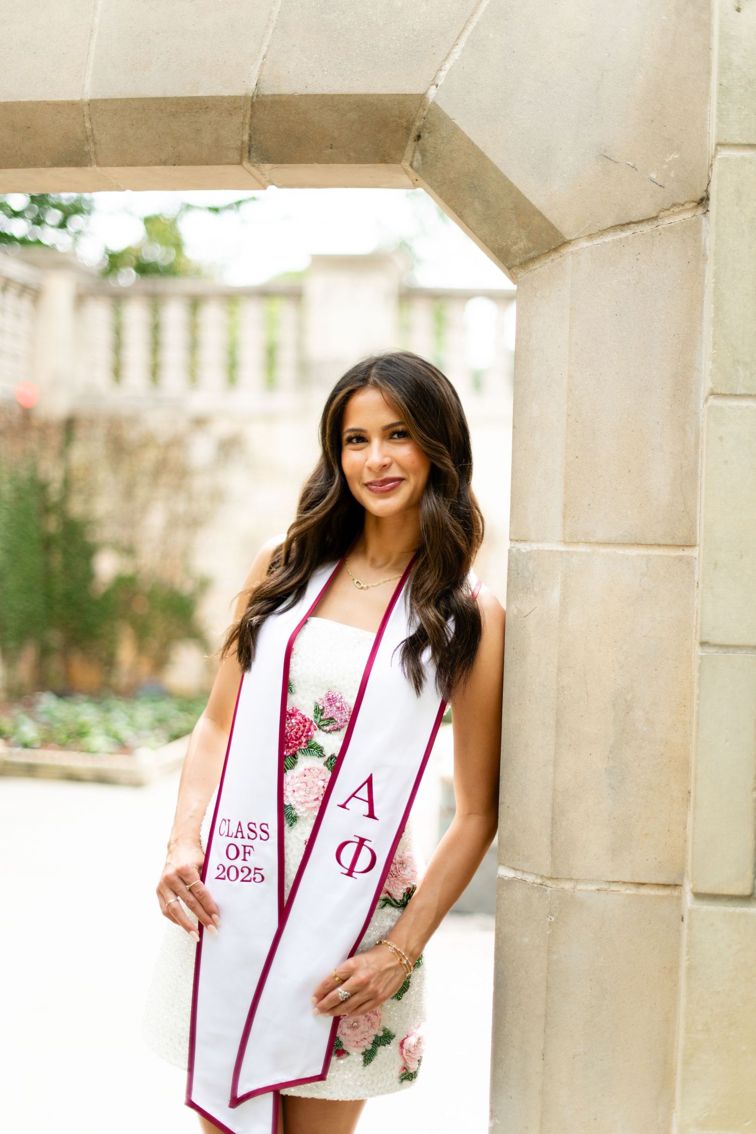 Graduate wearing white dress and red sorority stash poses under stone archway.