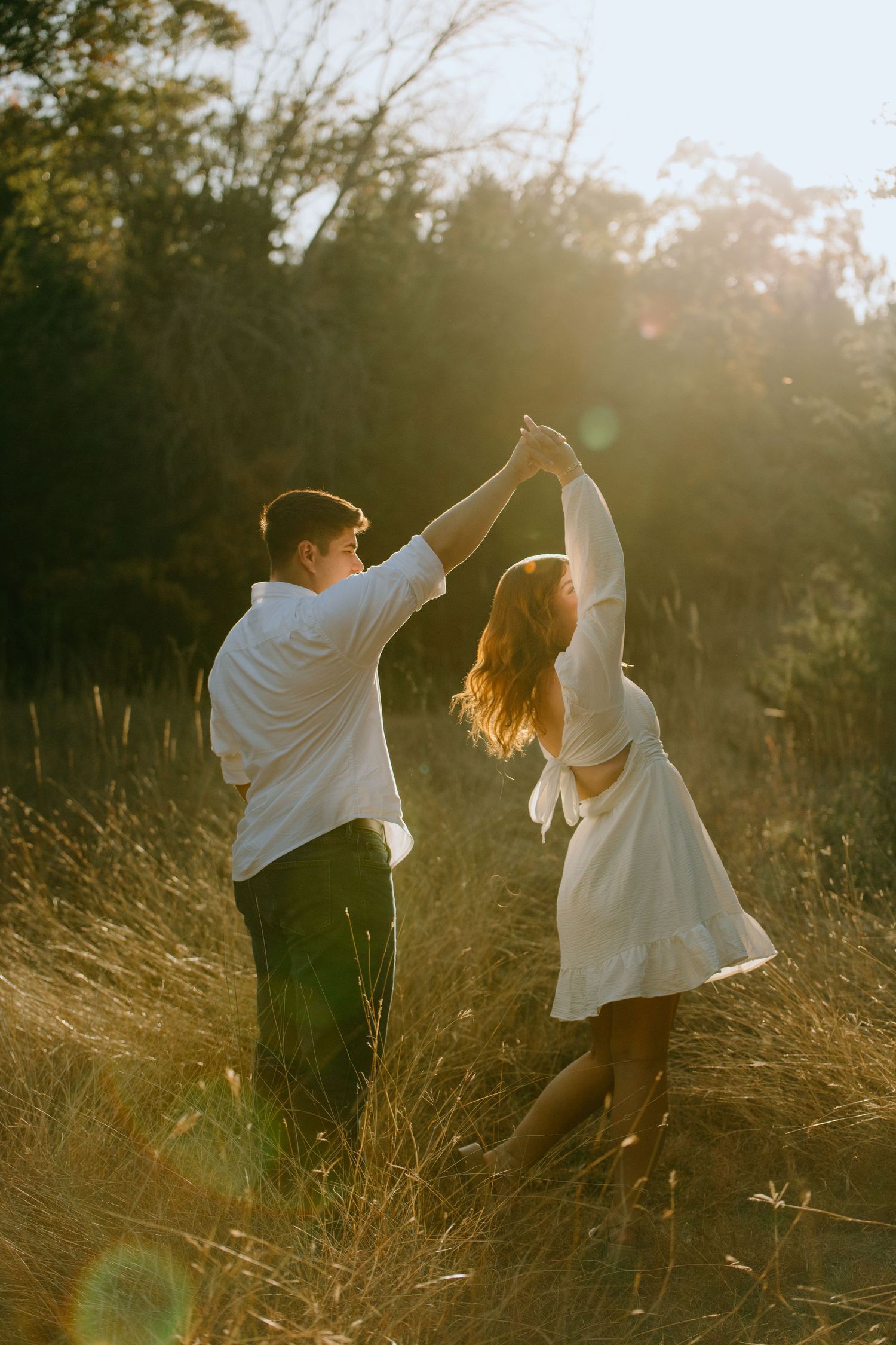 A couple dances together in a golden sunlit field of tall grass during sunset, creating a romantic silhouette.