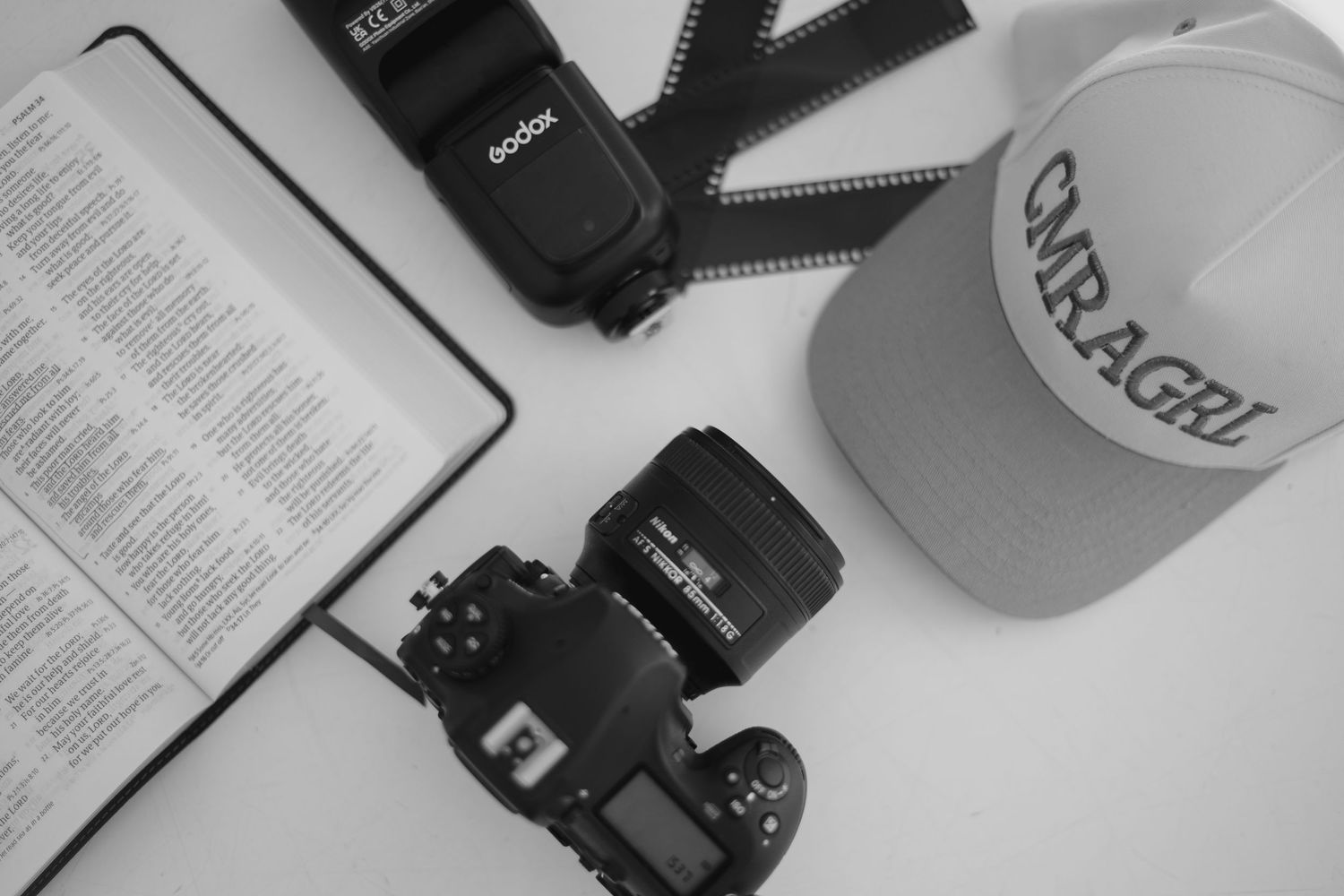 A black and white photo showing a DSLR camera, baseball cap, flash unit and open book arranged on a surface.