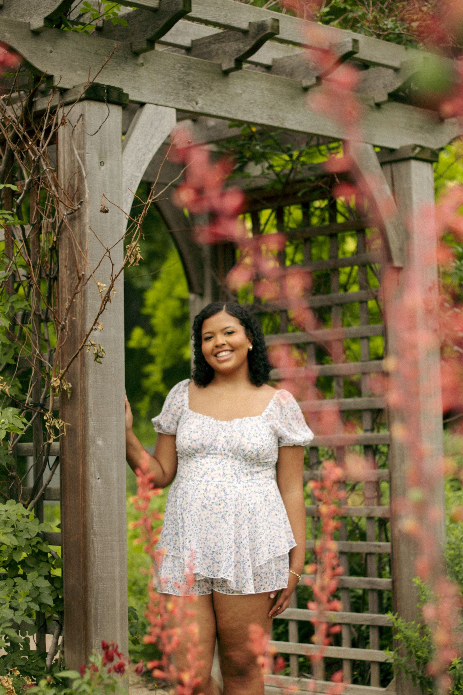 Woman in white lace dress poses under garden arbor with pink flowers.