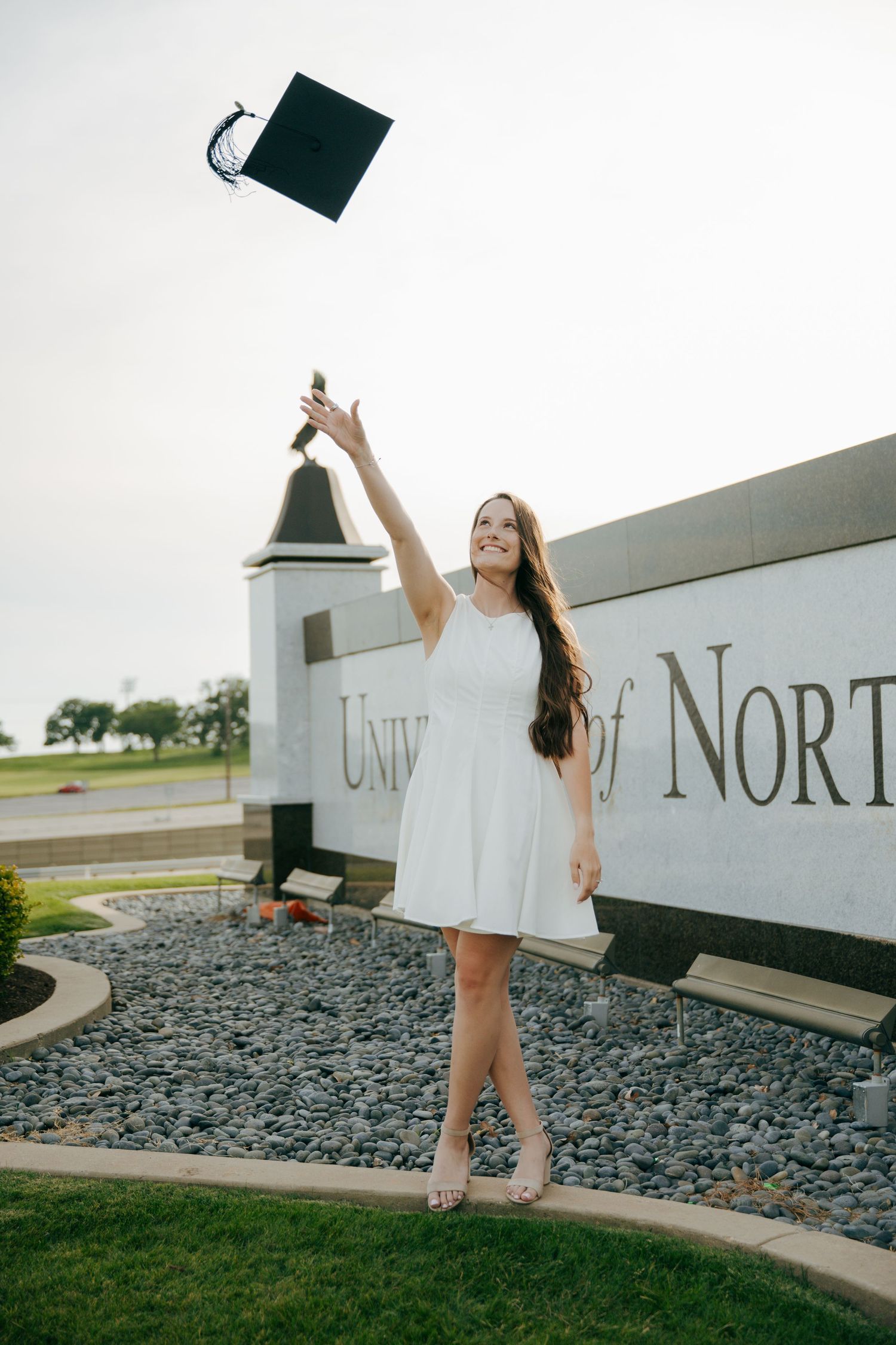 A smiling graduate in a white dress tosses her cap in front of a University of North Texas sign.