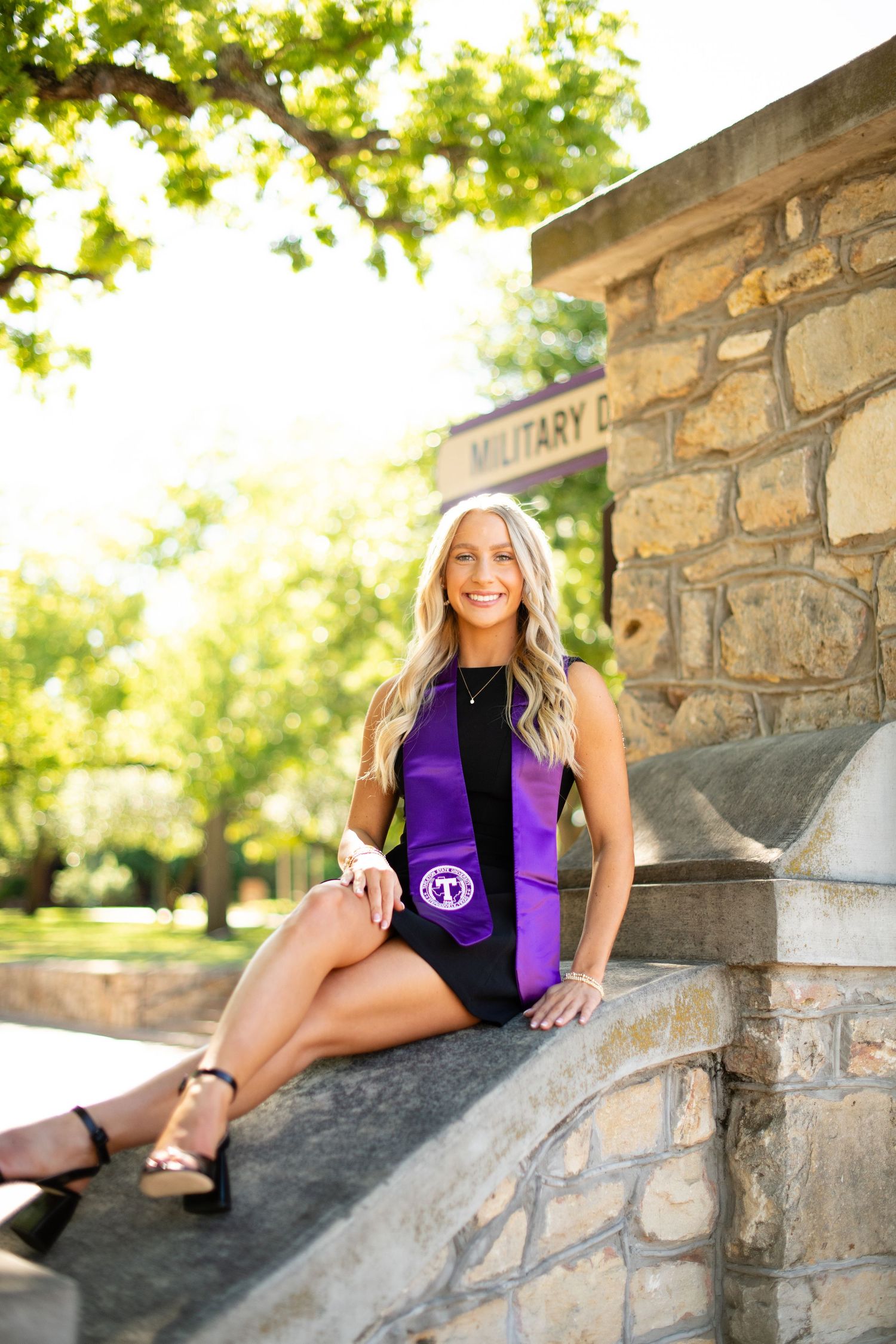 Graduate in black dress and purple stole poses on stone steps in sunny outdoor setting.