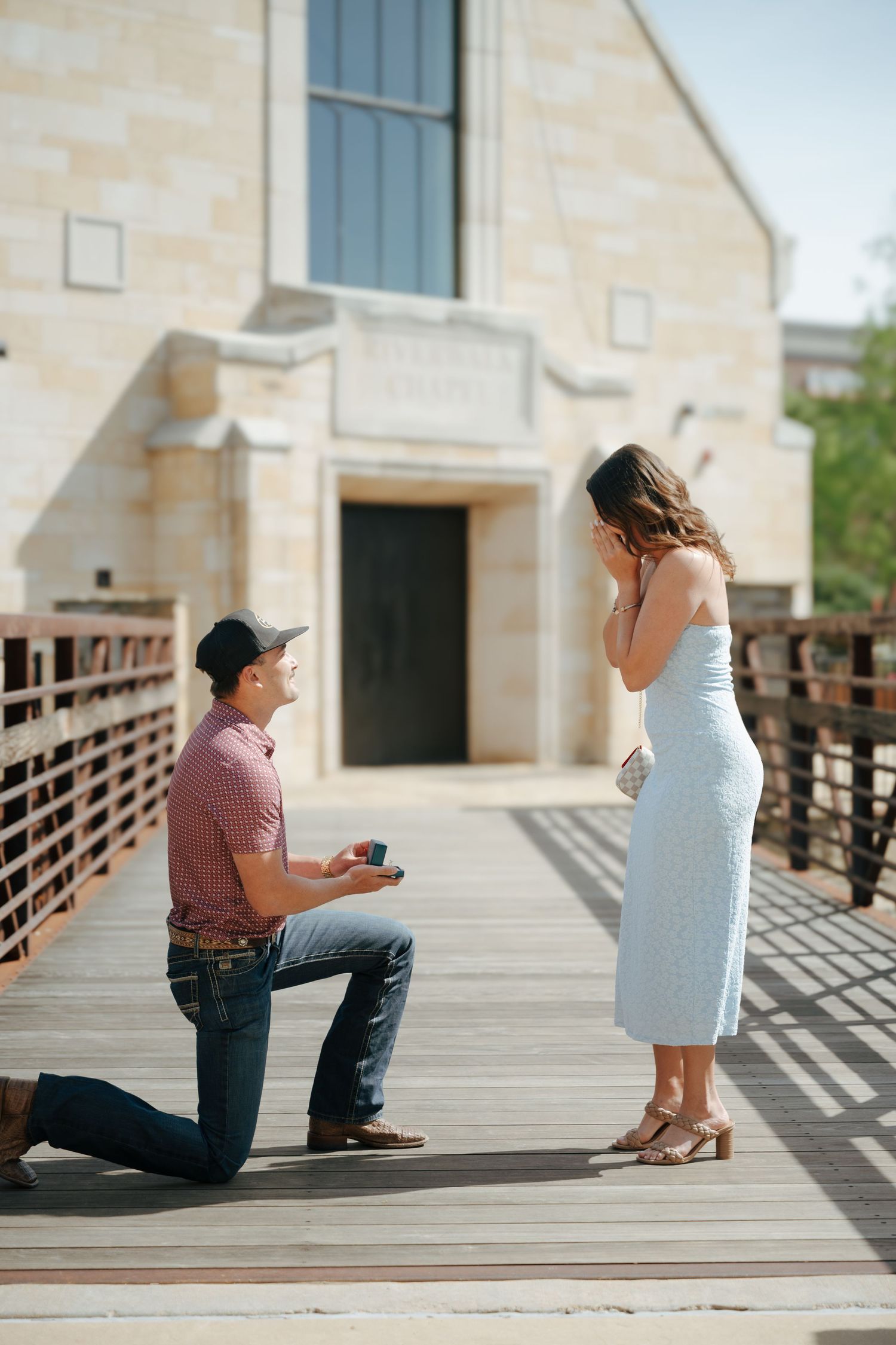 Man kneeling on wooden bridge proposing to woman in light blue dress outside stone church building.