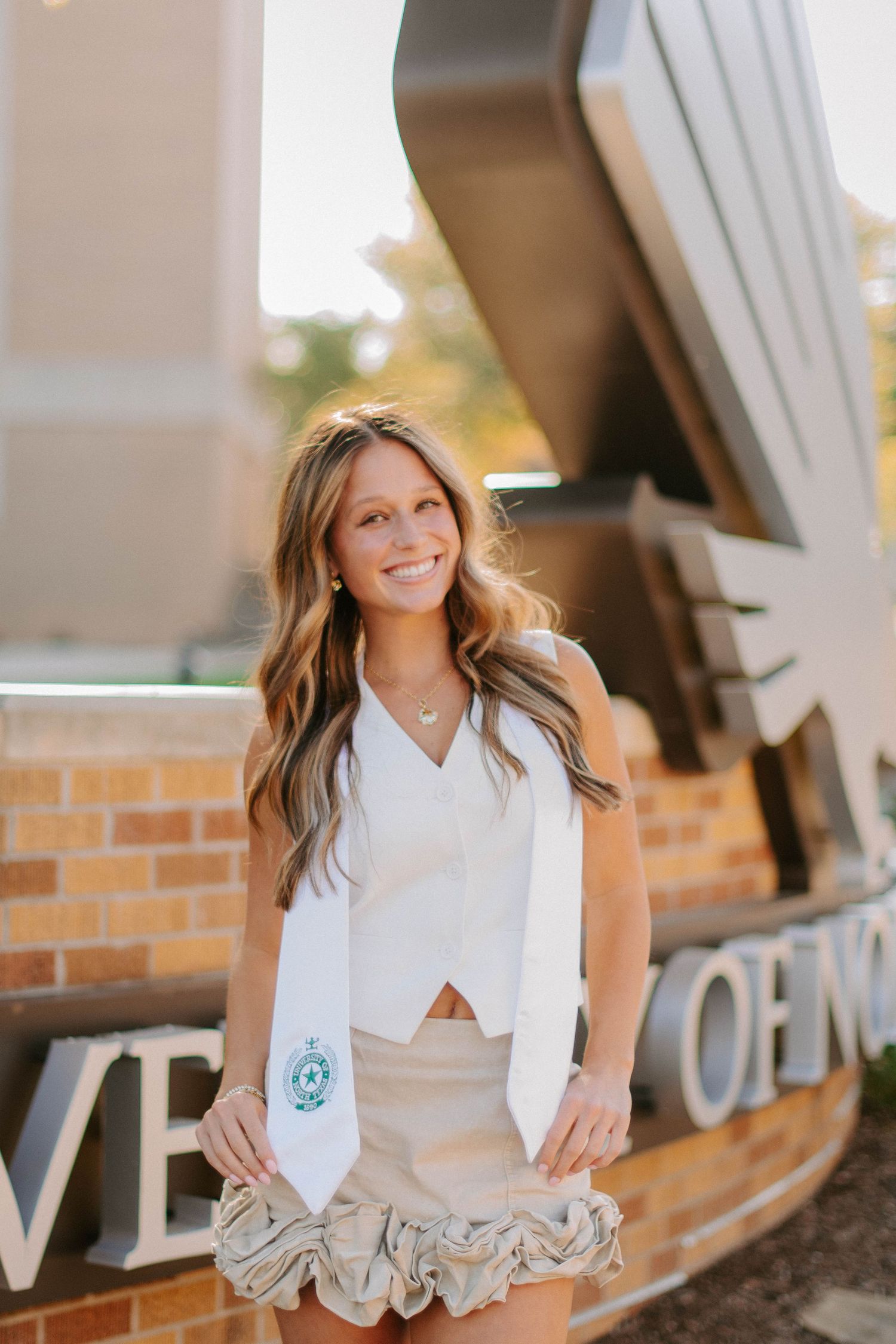 Graduate in white dress and stole stands confidently near university signage.
