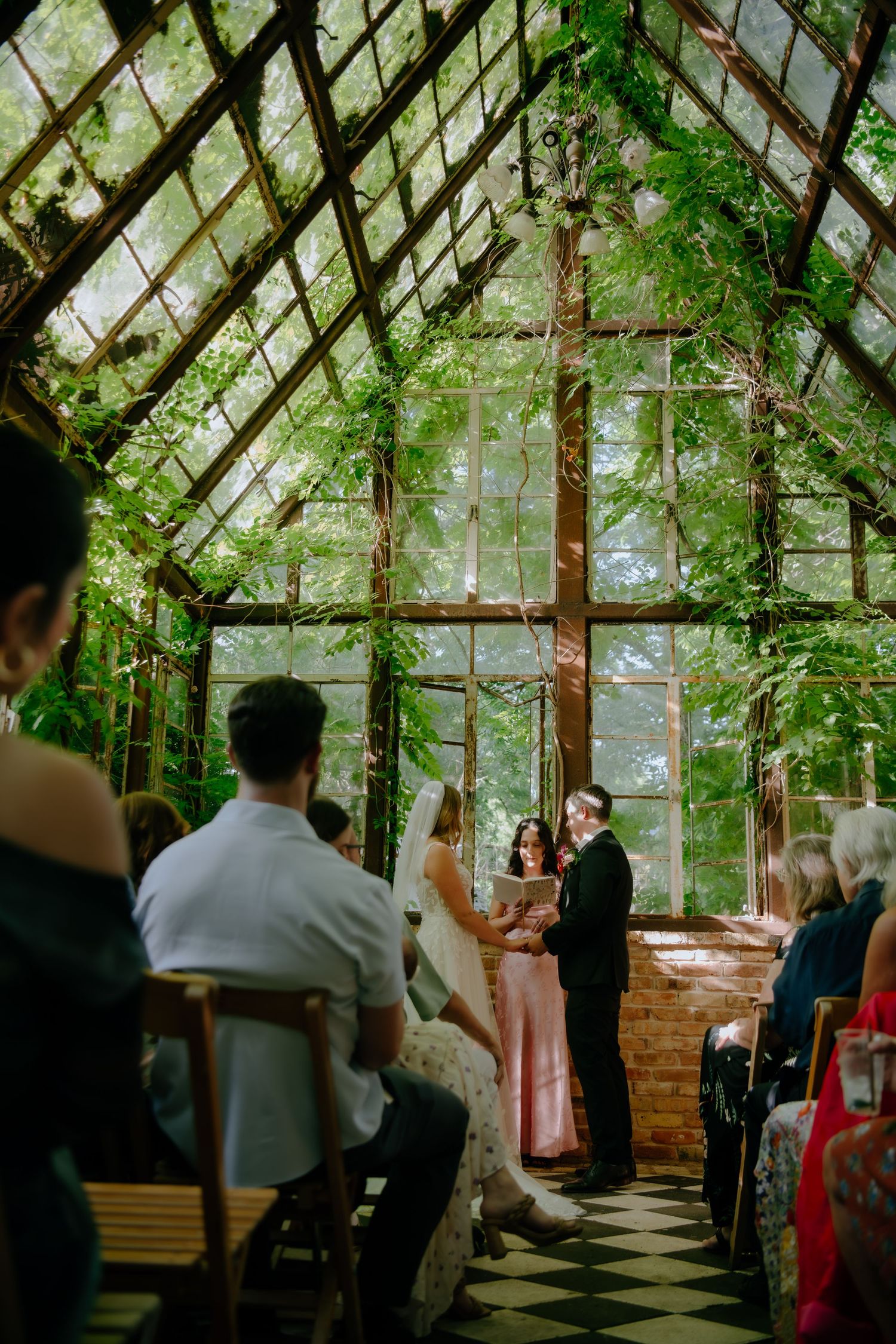 Wide view of a greenhouse wedding venue with guests seated on wooden chairs beneath glass ceiling.