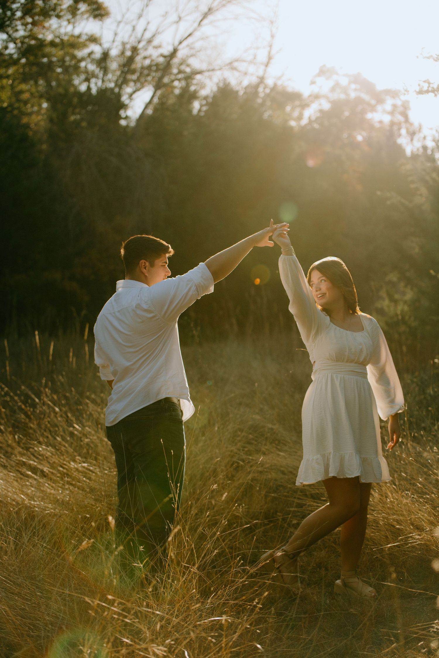 A couple dances playfully in a sunlit meadow with tall golden grass at sunset.
