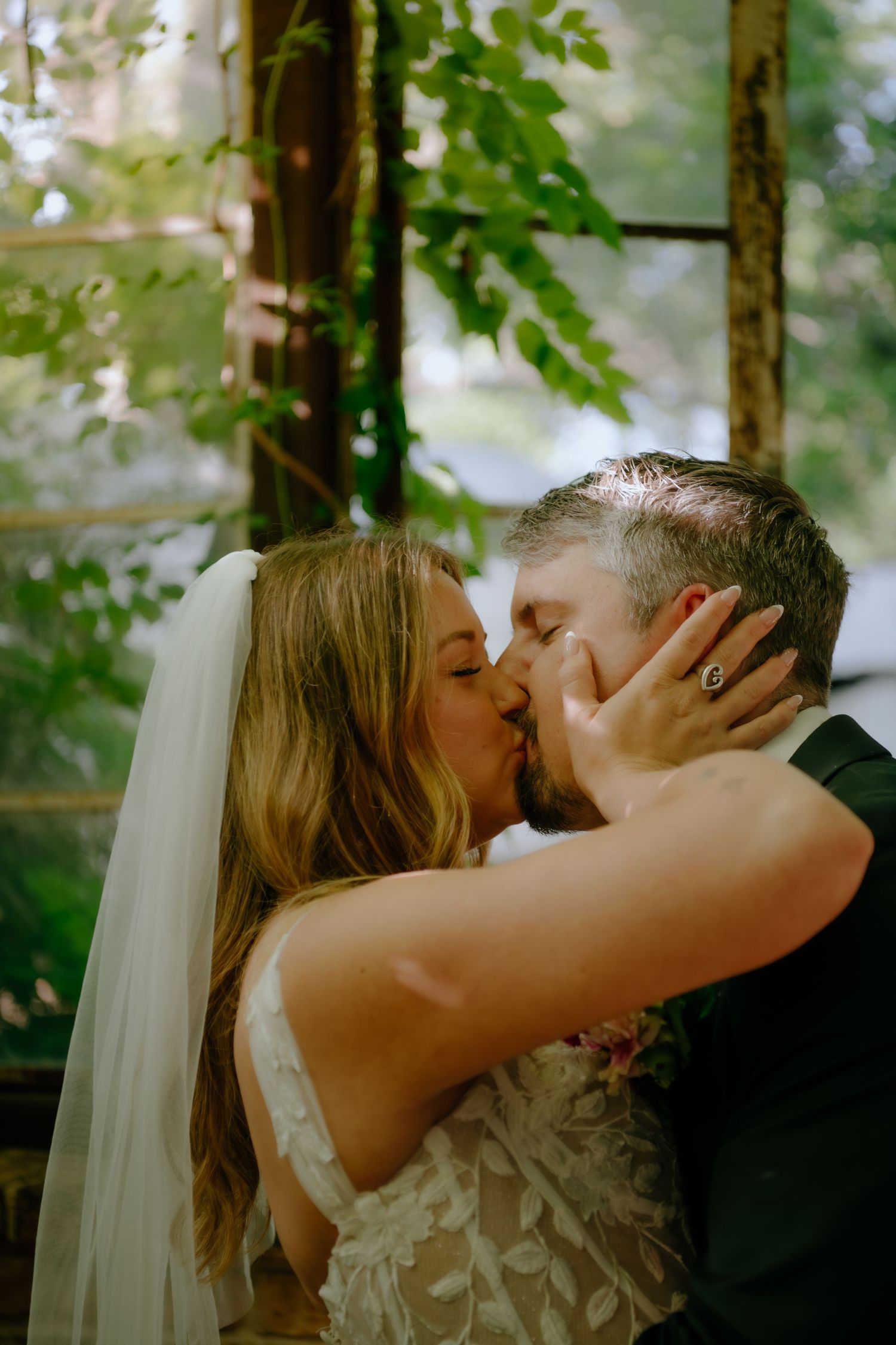 A romantic wedding kiss in a sunlit greenhouse filled with vines and natural light.