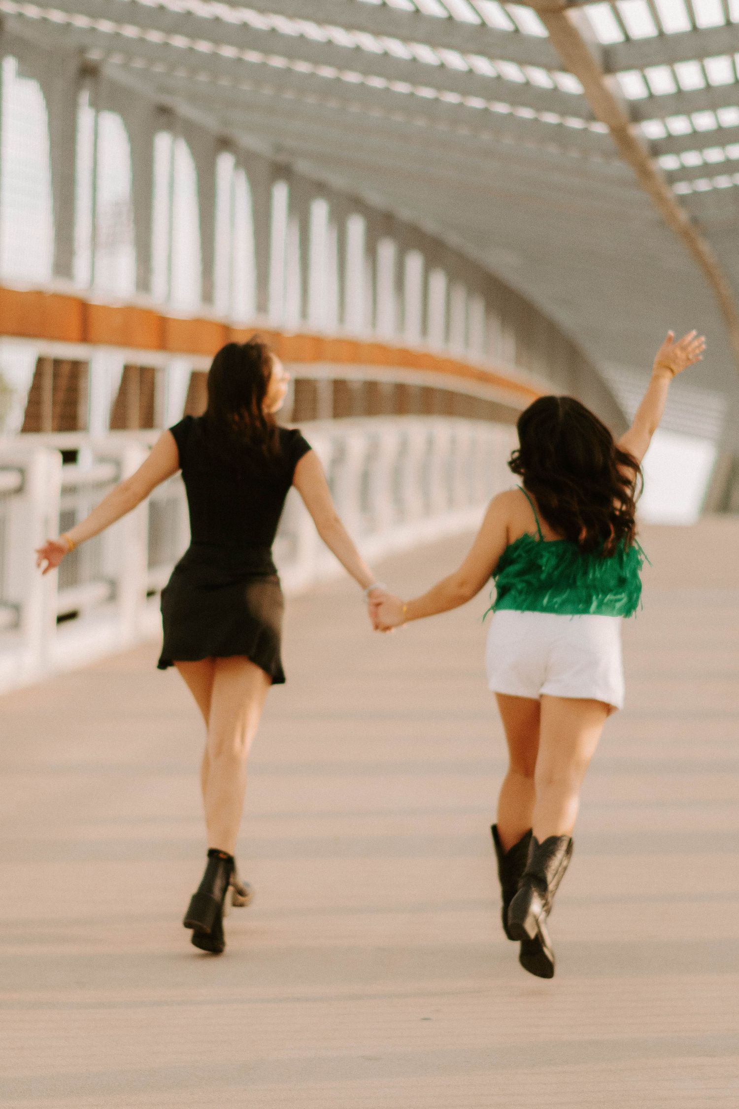 Two friends holding hands and running down a covered walkway wearing boots and summer outfits.