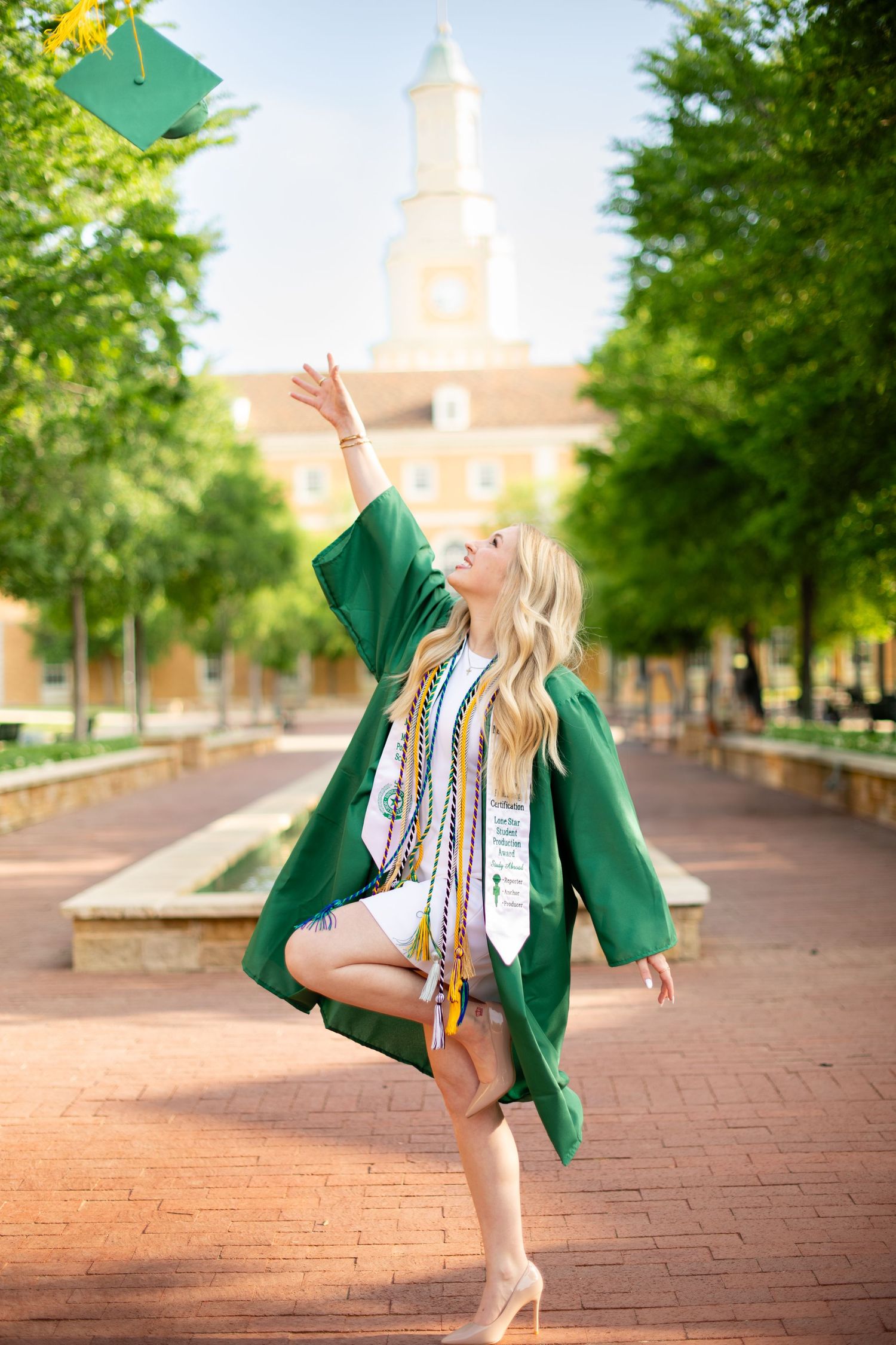 Graduate in green cap and gown celebrates on brick pathway with university building in background.