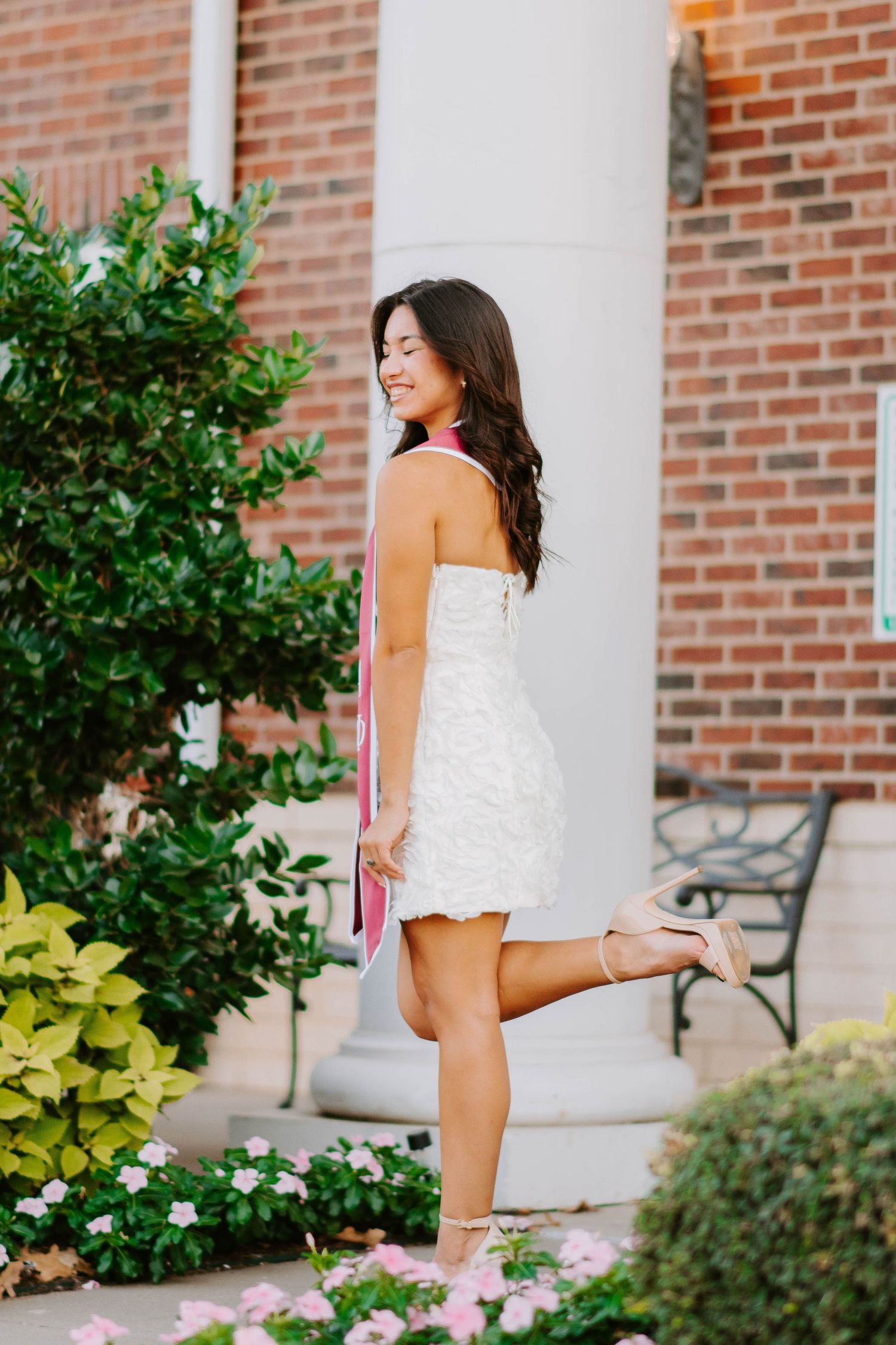 White lace dress poses elegantly against brick column surrounded by garden flowers.