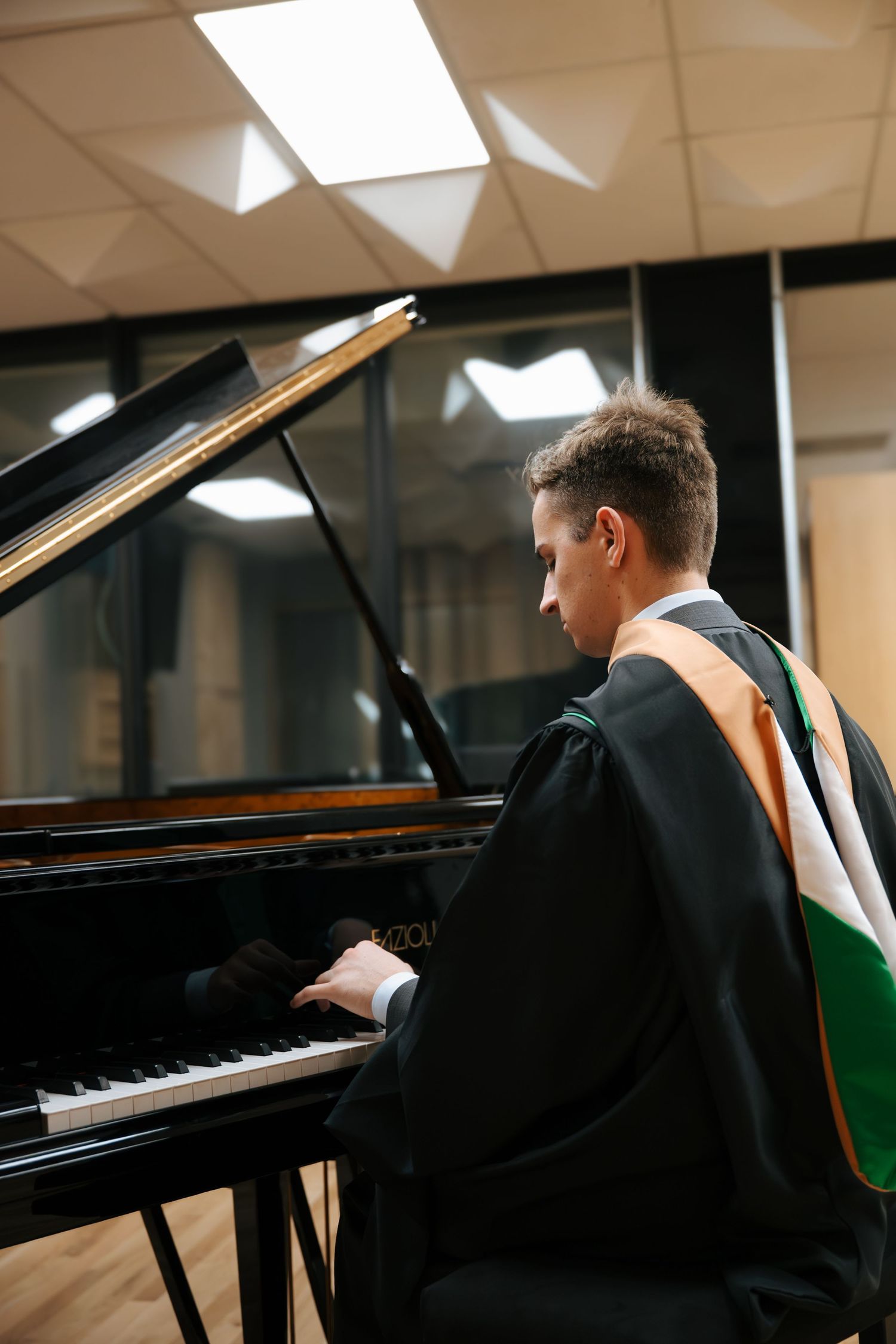 Graduate in cap and gown playing a grand piano in a music studio during a graduation ceremony.