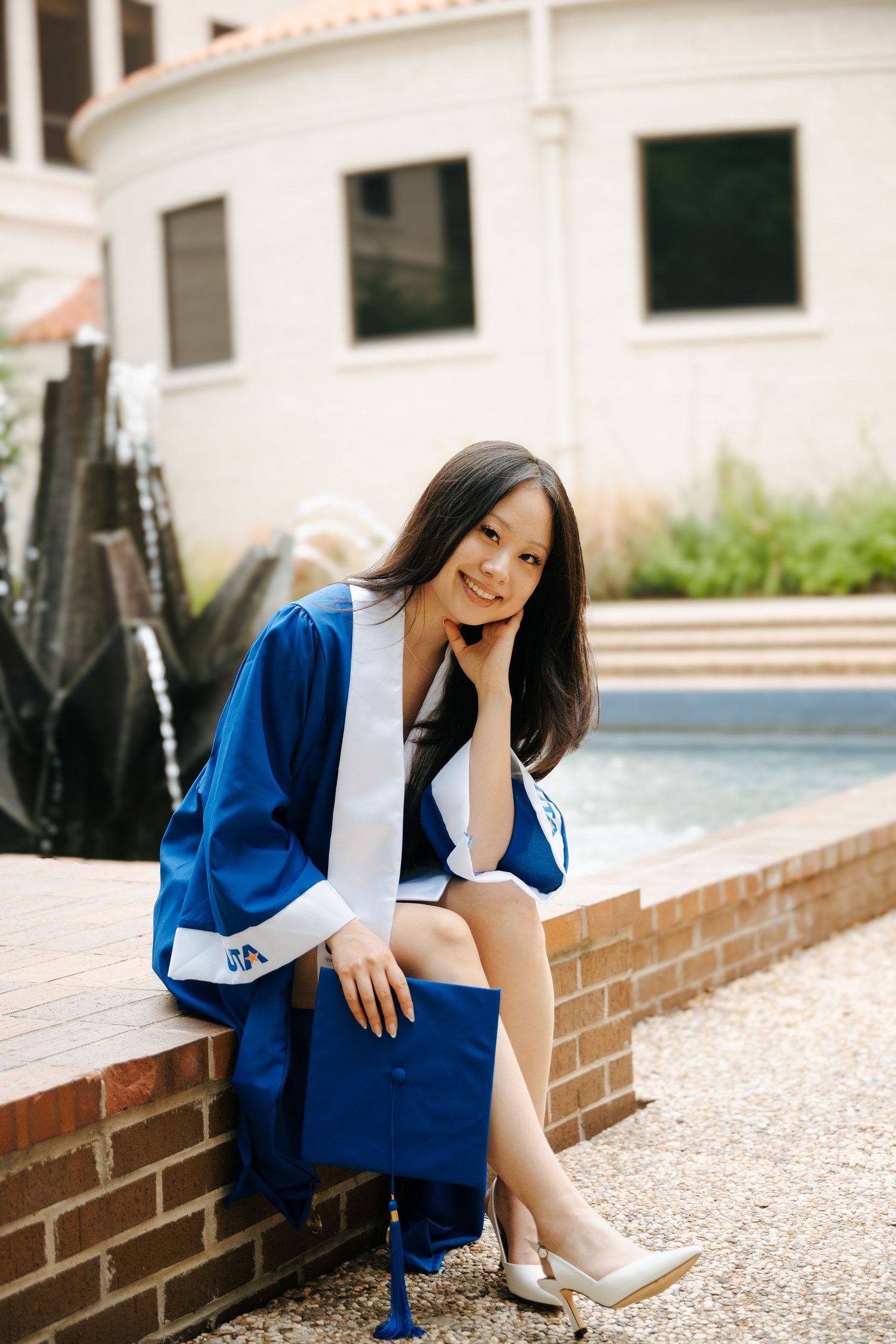 Young woman in blue graduation gown sitting on brick ledge, smiling, holding cap, wearing heels outdoors.