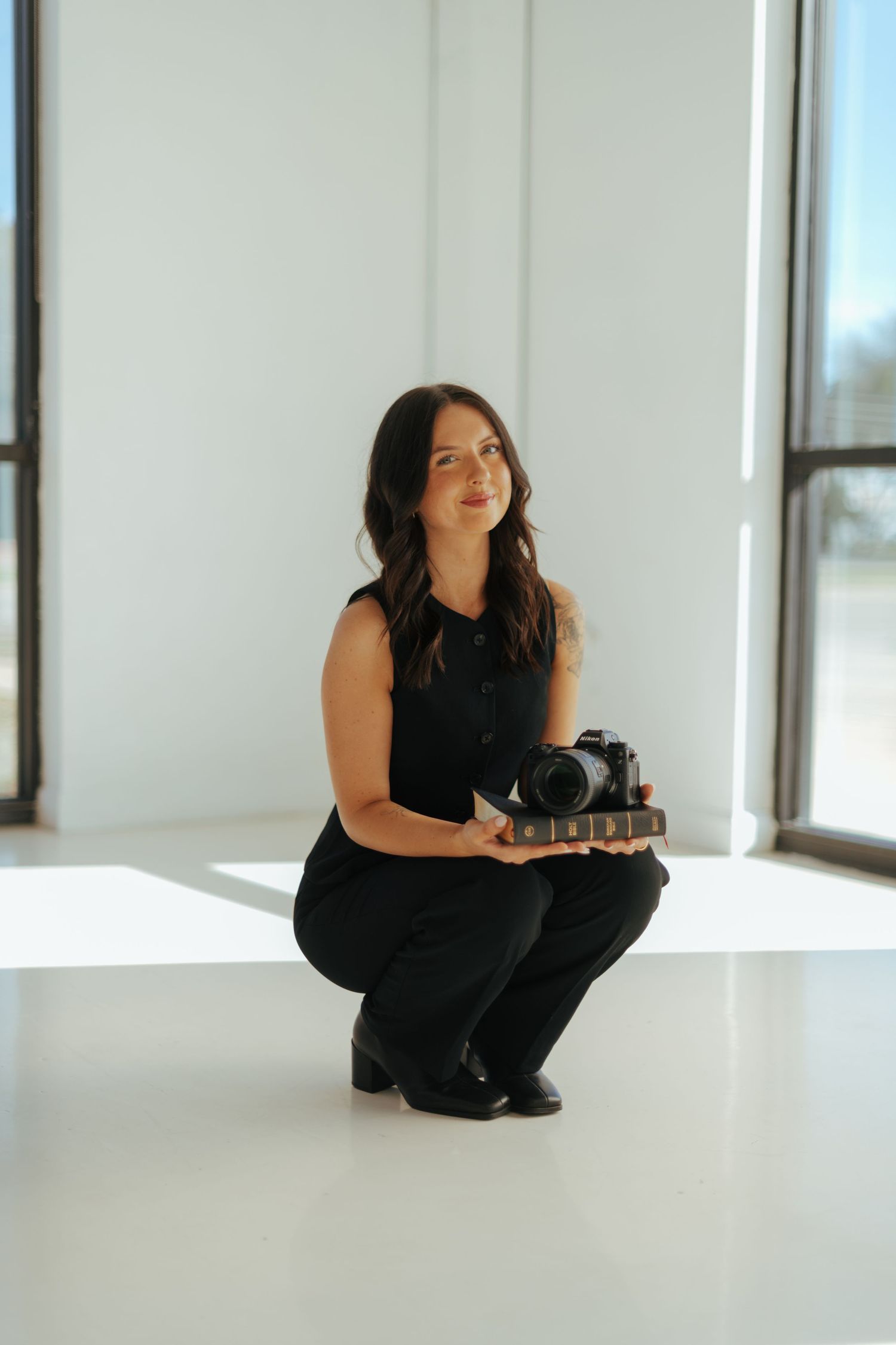 Professional photographer in black outfit sits on white floor with camera in bright studio space.