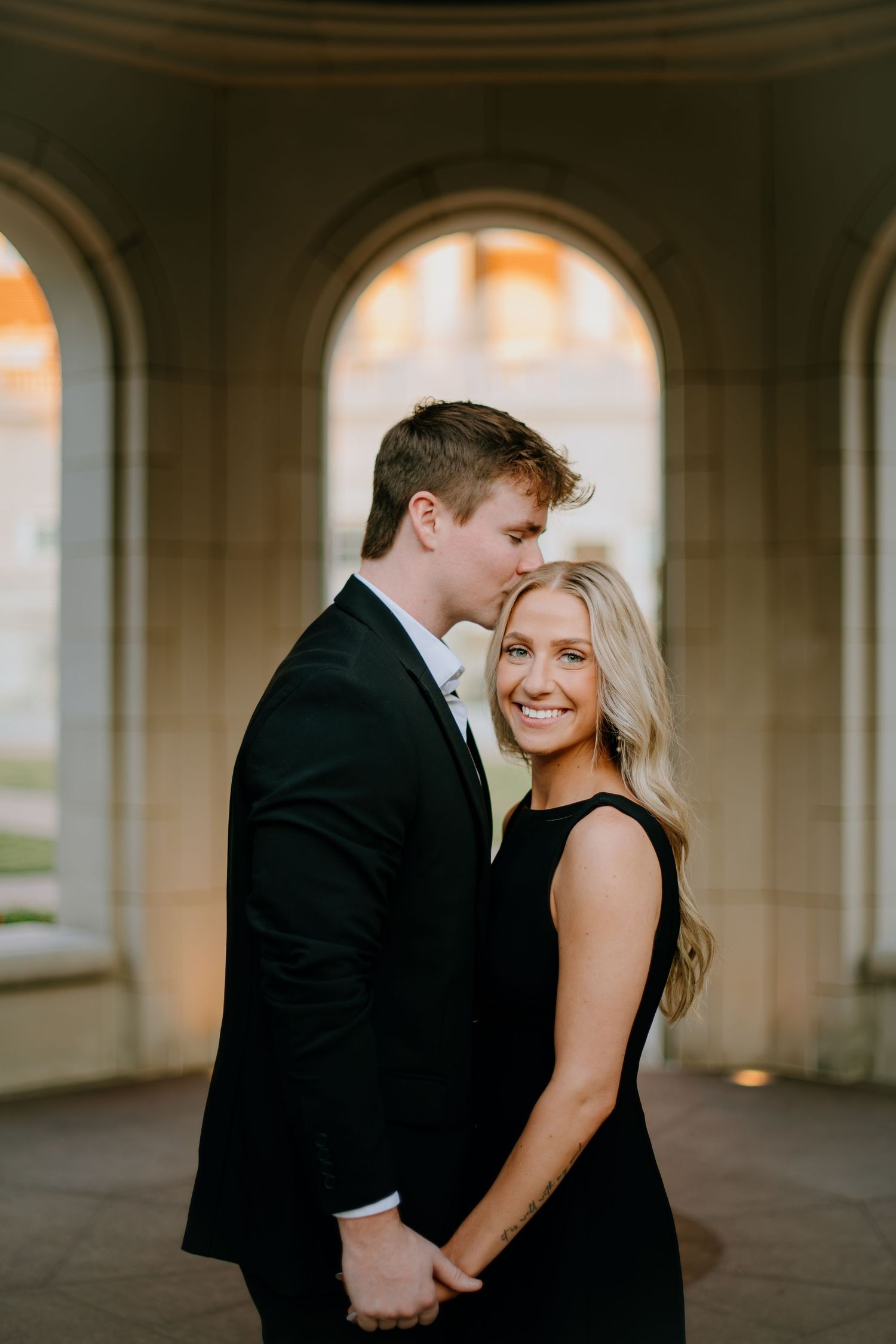 A romantic couple in formal attire embraces under an arched passageway during a wedding photoshoot.