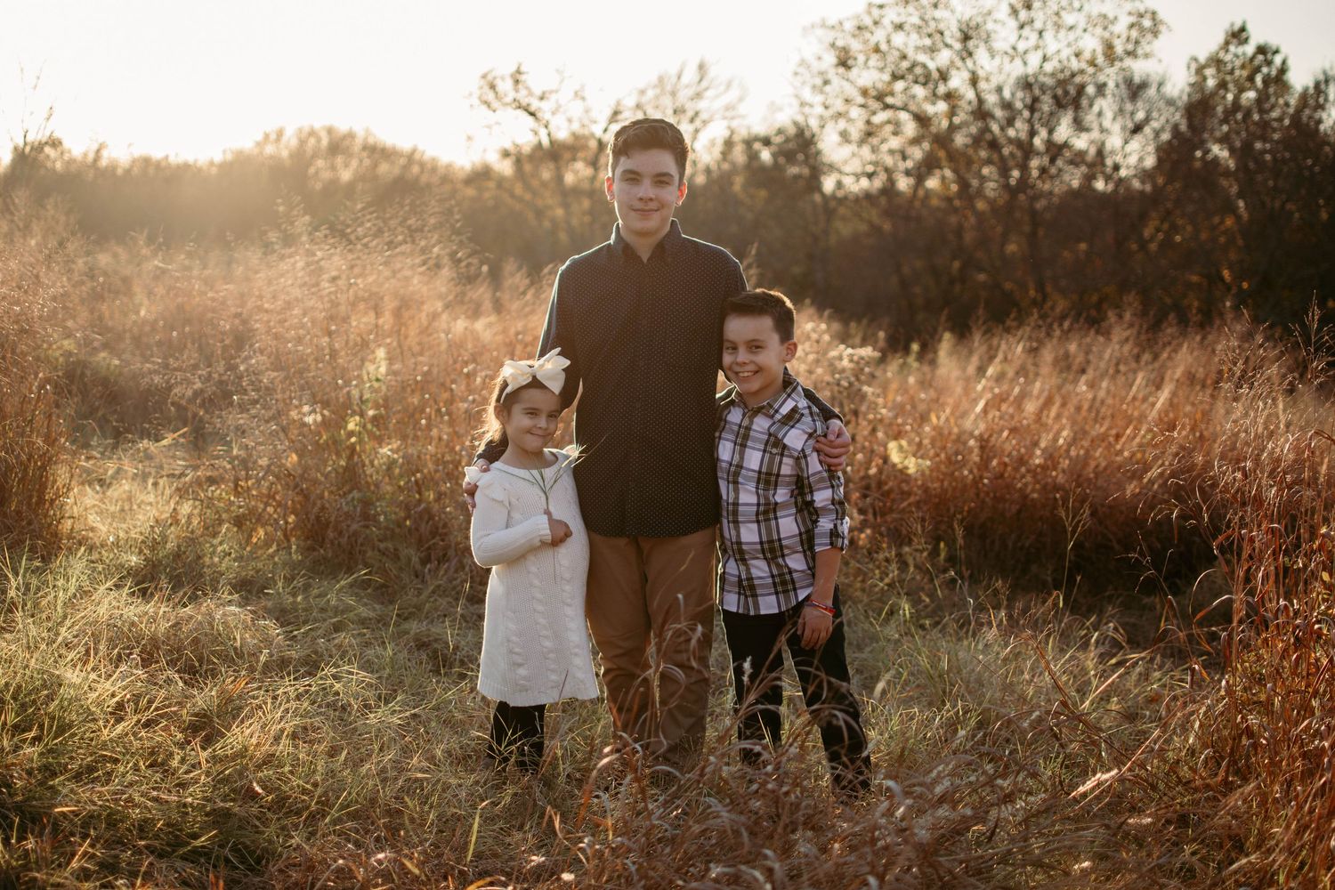 Three people posing together in a scenic field during golden hour with natural grass and foliage.