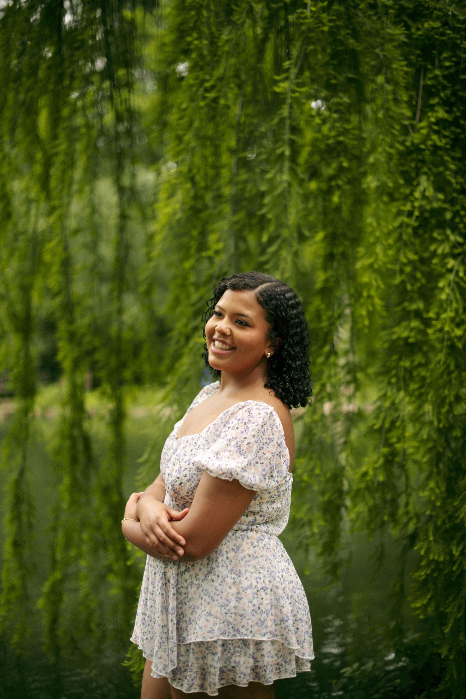 Woman in white floral dress poses under weeping willow tree branches near water.