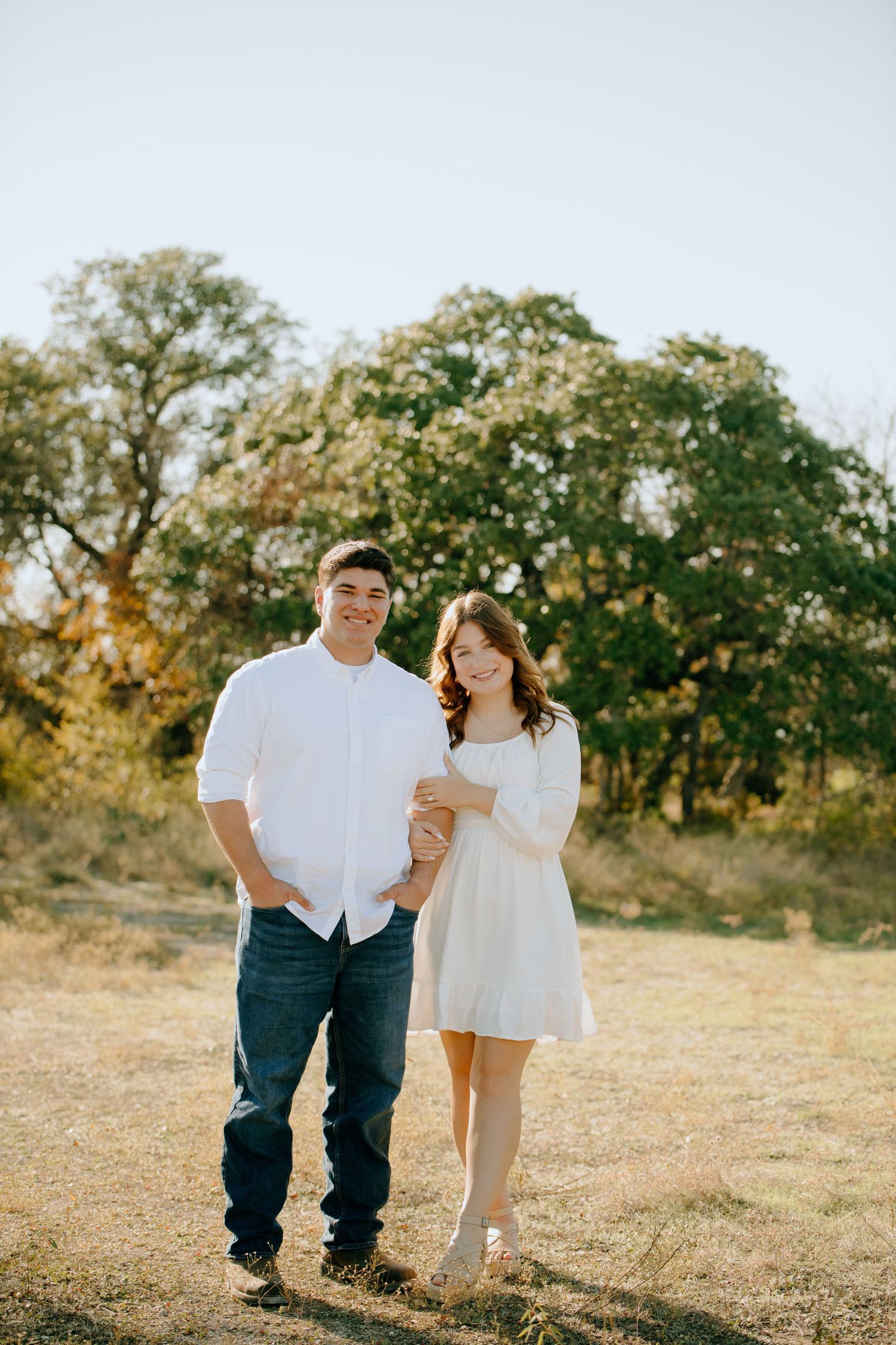 A couple walking together in a sunny field with blooming trees in the background.