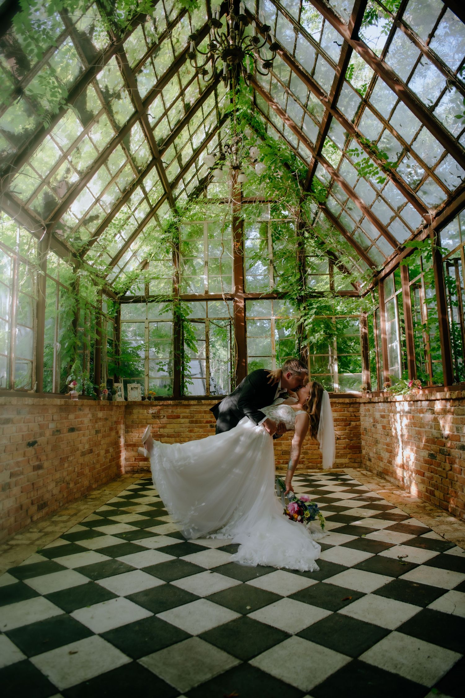 Dramatic wedding dip pose on checkered floor in a greenhouse with vaulted glass ceiling.