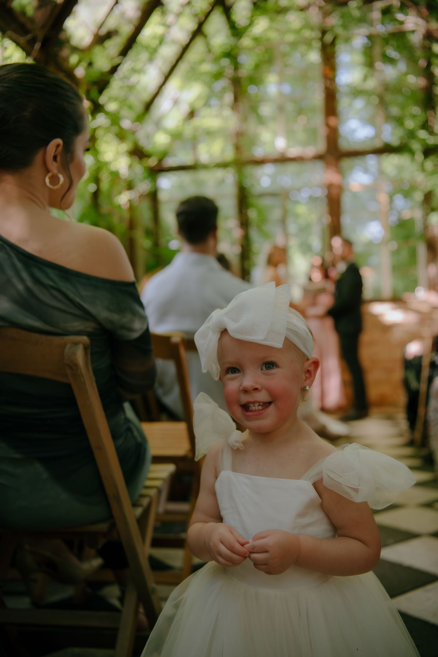 A young child in a white dress sits among wedding guests during a ceremony.