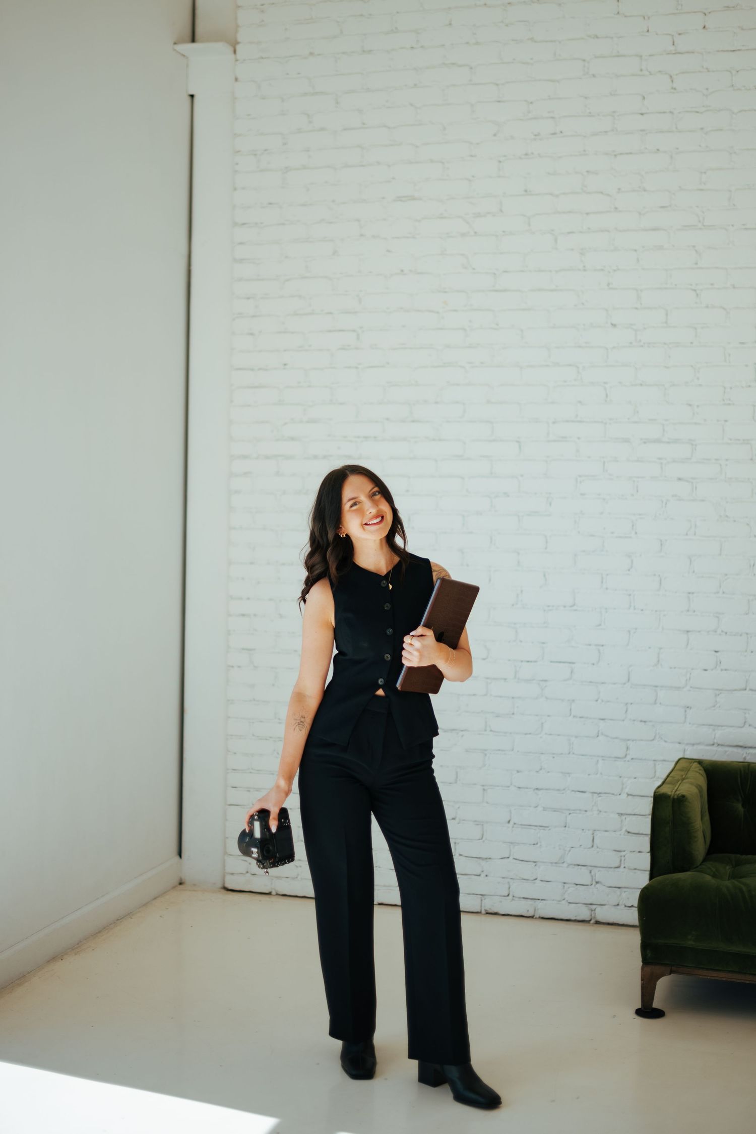Professional wearing an all-black business outfit stands confidently against white brick wall holding notebook and heels.