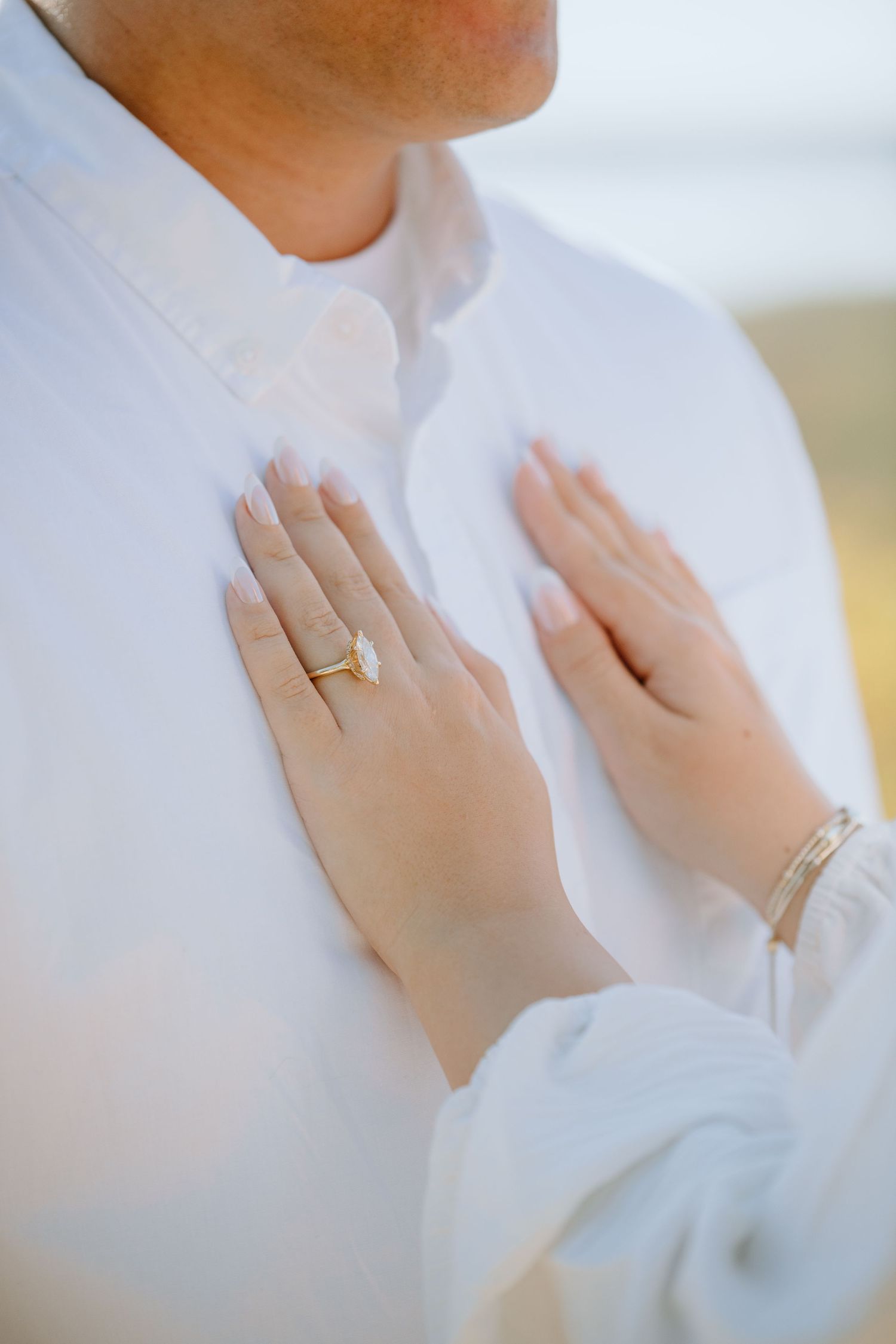 Close-up of hands wearing a gold wedding ring and bracelet against a white dress in soft natural light.