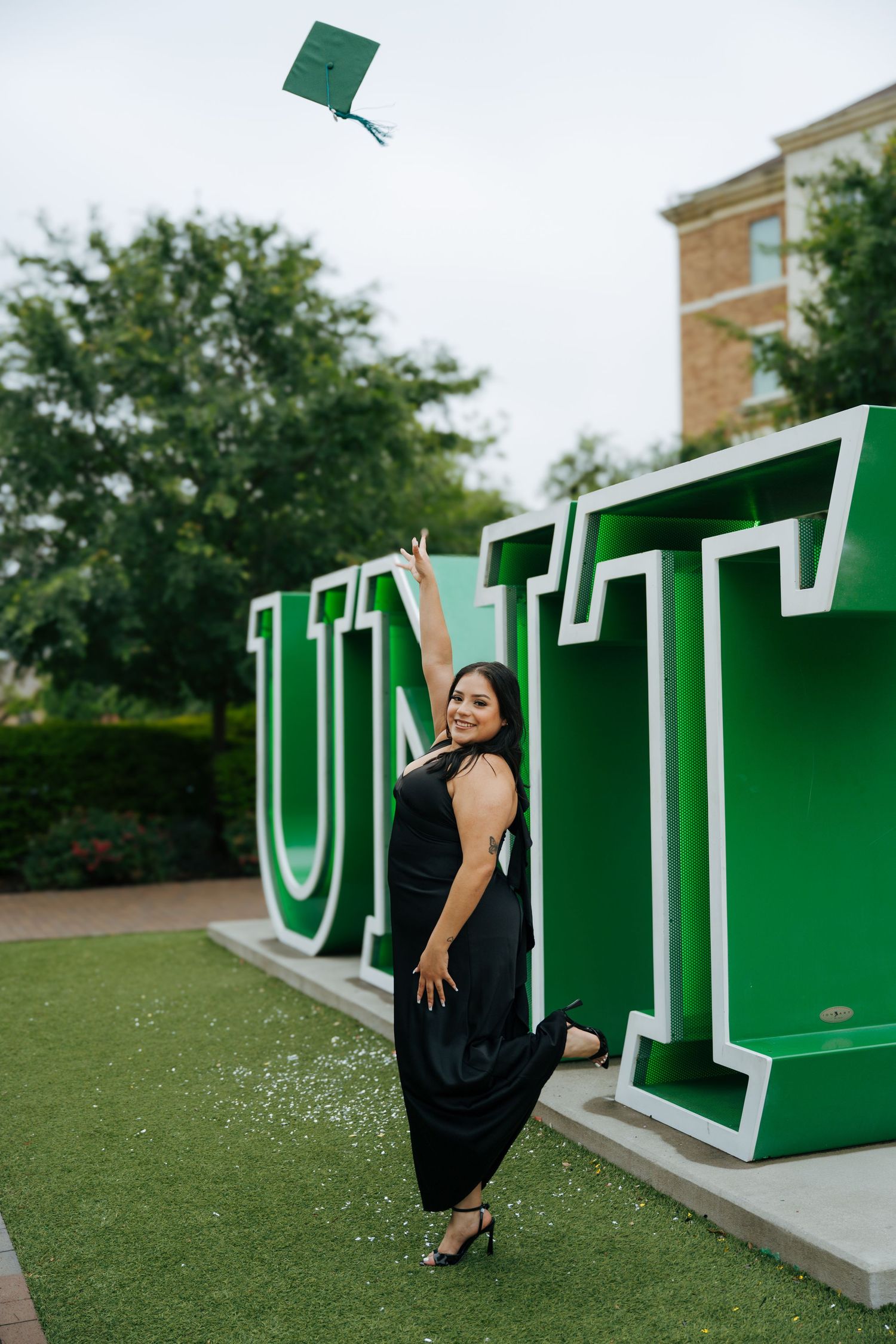 A woman in a black dress tosses her green graduation cap in front of large UNT letters on a university campus.