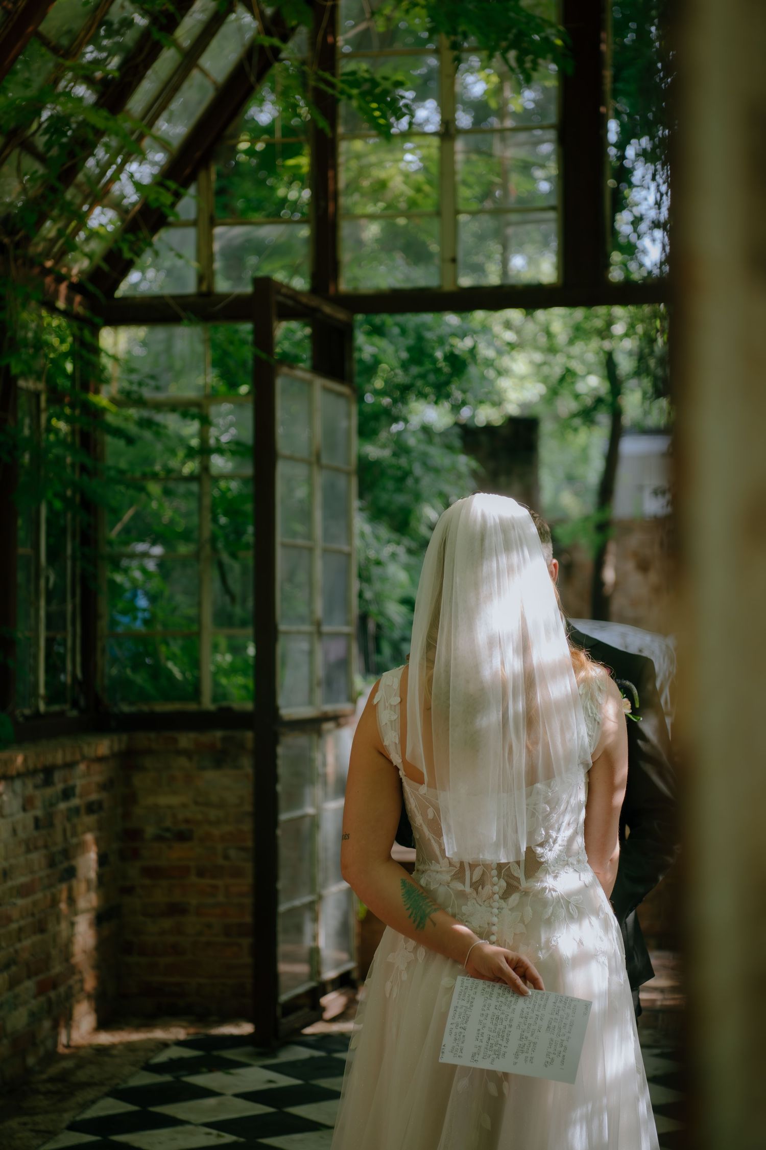 Bride in white gown and veil stands in a vintage greenhouse doorway surrounded by lush green foliage.