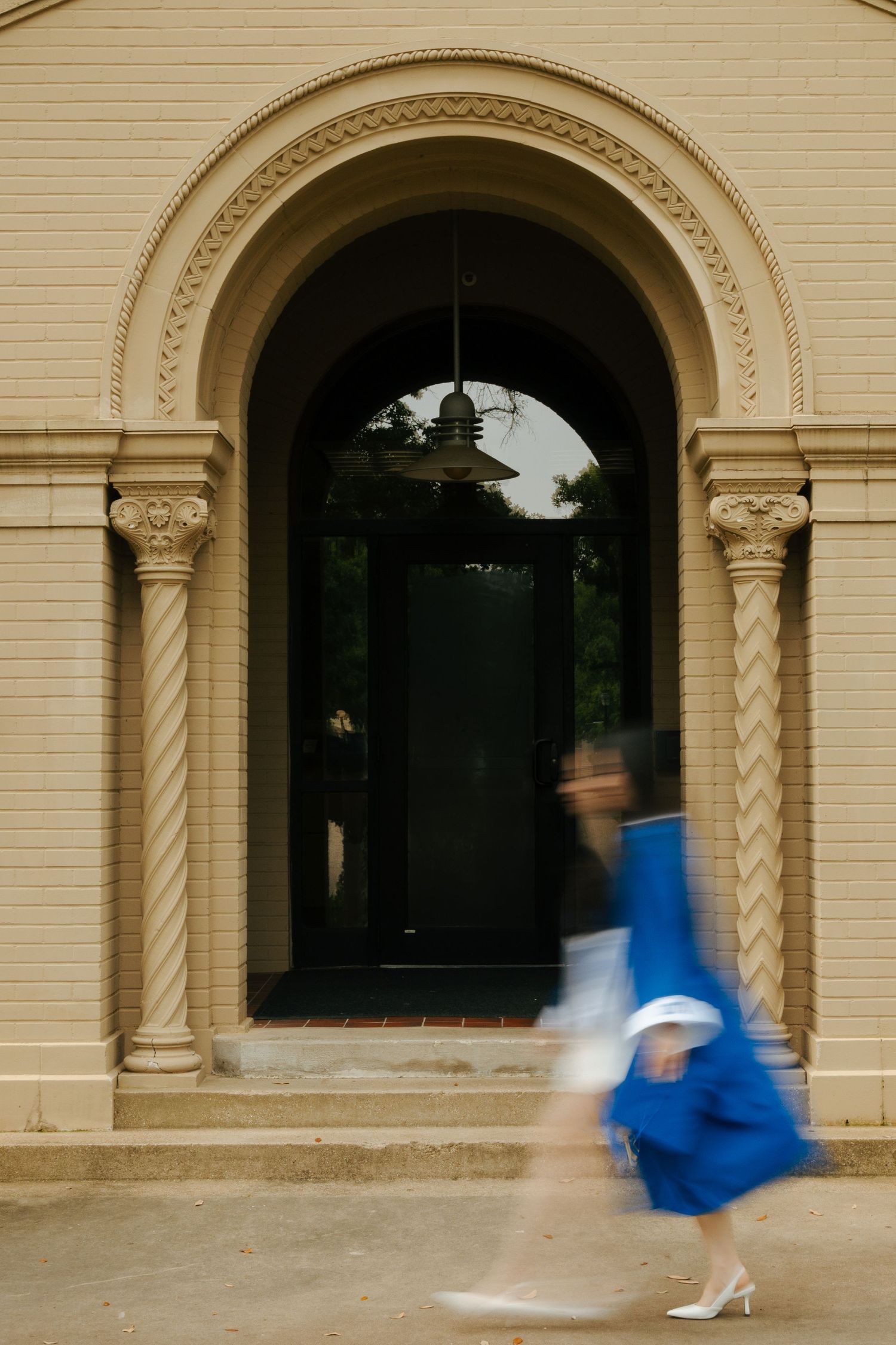 A blurred graduate in blue cap and gown walks past an ornate arched doorway of a beige stone building.