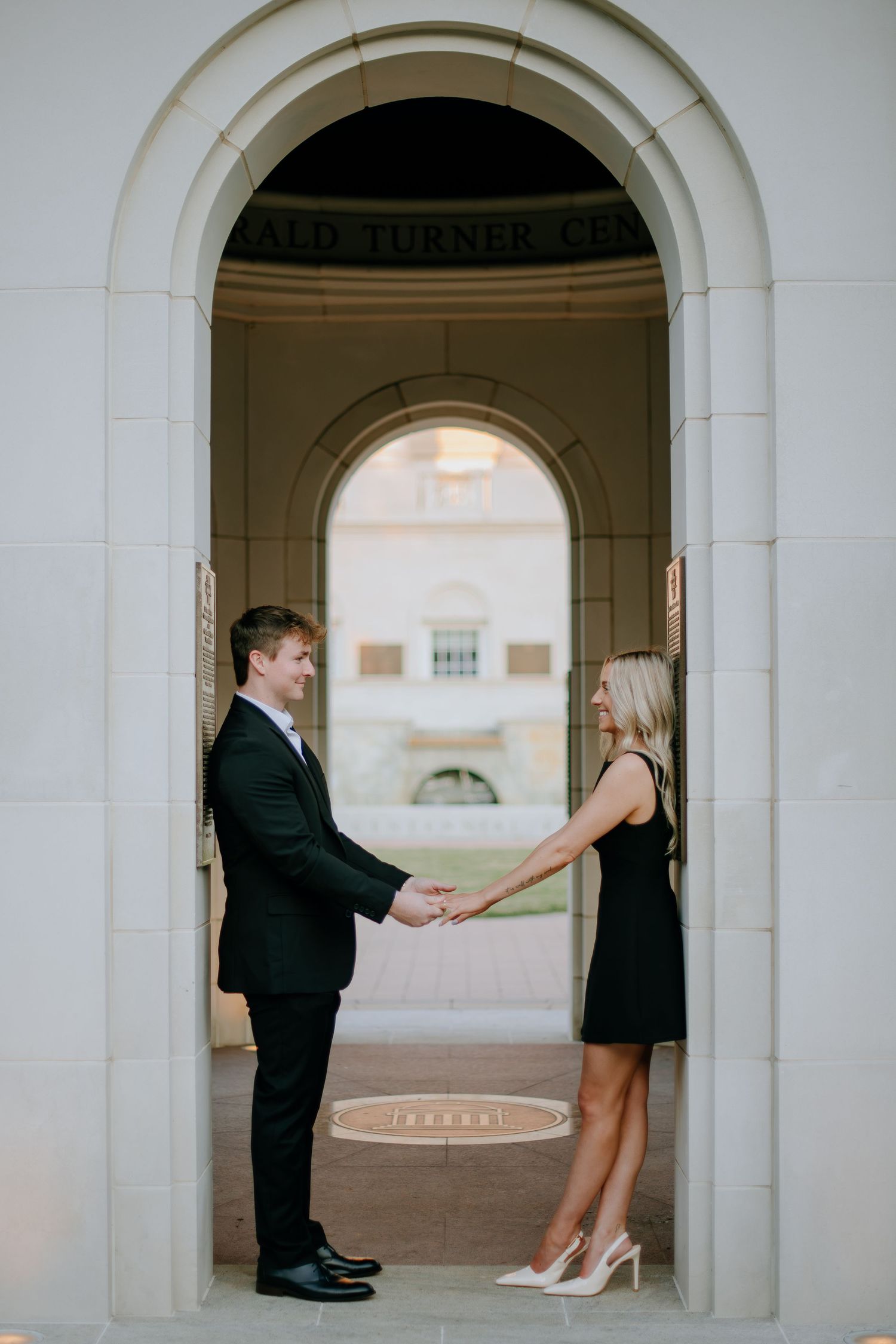 A romantic couple shares playful moments in an elegant white archway corridor.