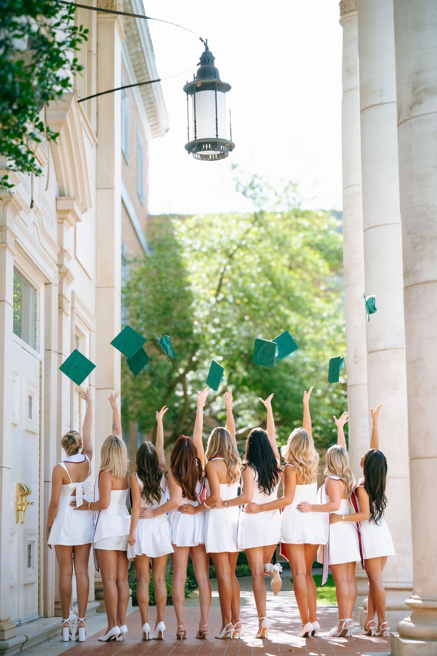 Group of female graduates in white dresses tossing green graduation caps in the air outside a classical building.
