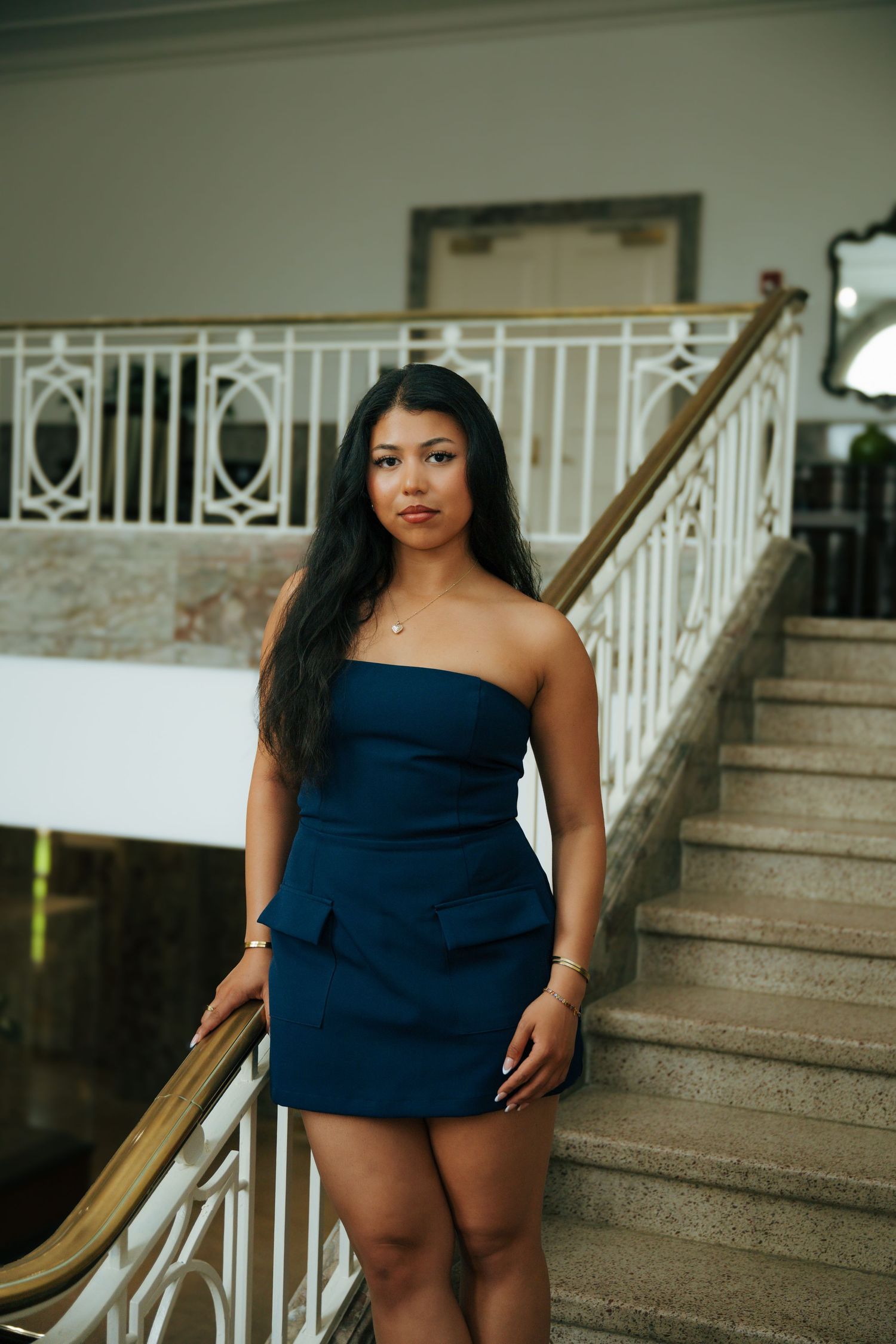 Young woman in navy strapless mini dress posing elegantly on marble staircase with white decorative railings.