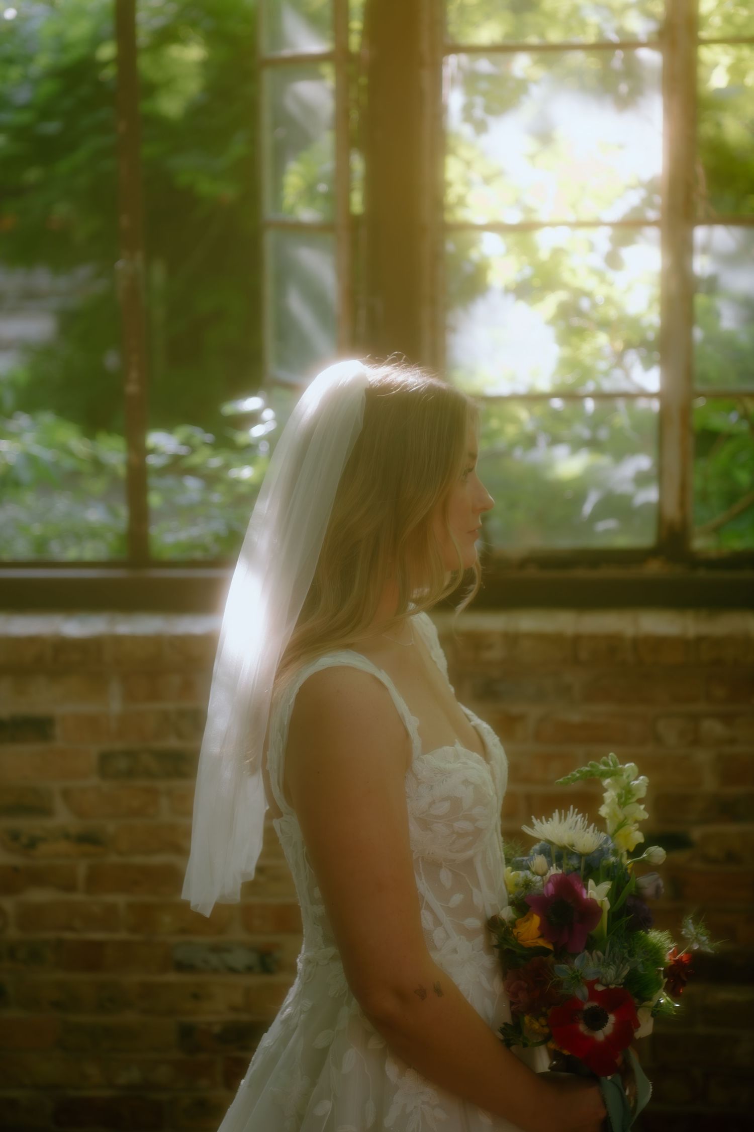 Ethereal bridal portrait in a sunlit greenhouse with dramatic window light and floral bouquet.