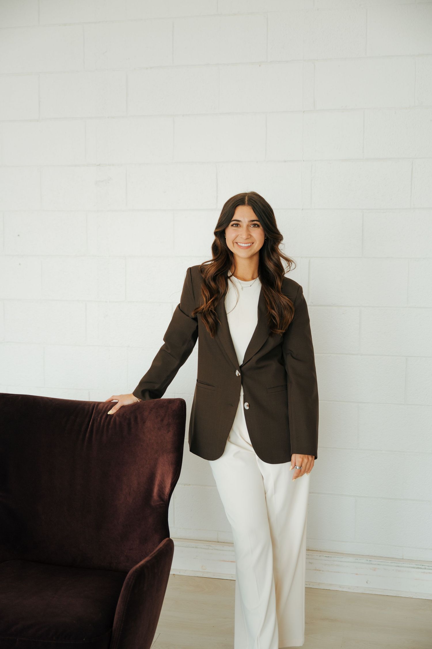 A young woman in a brown blazer and white pants smiles while standing beside a dark velvet sofa against a white brick wall.