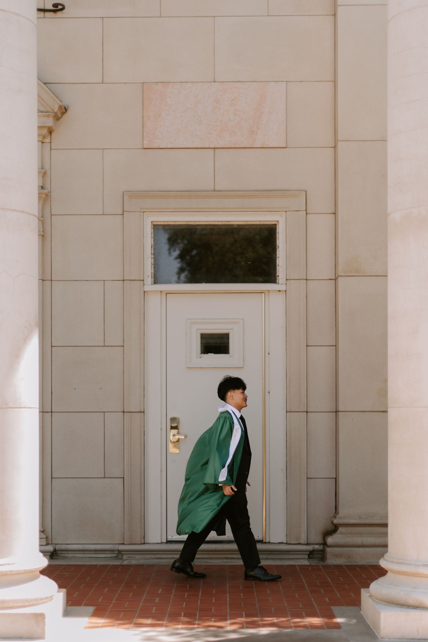 Graduate in green cap and gown poses confidently by white stone doorway.