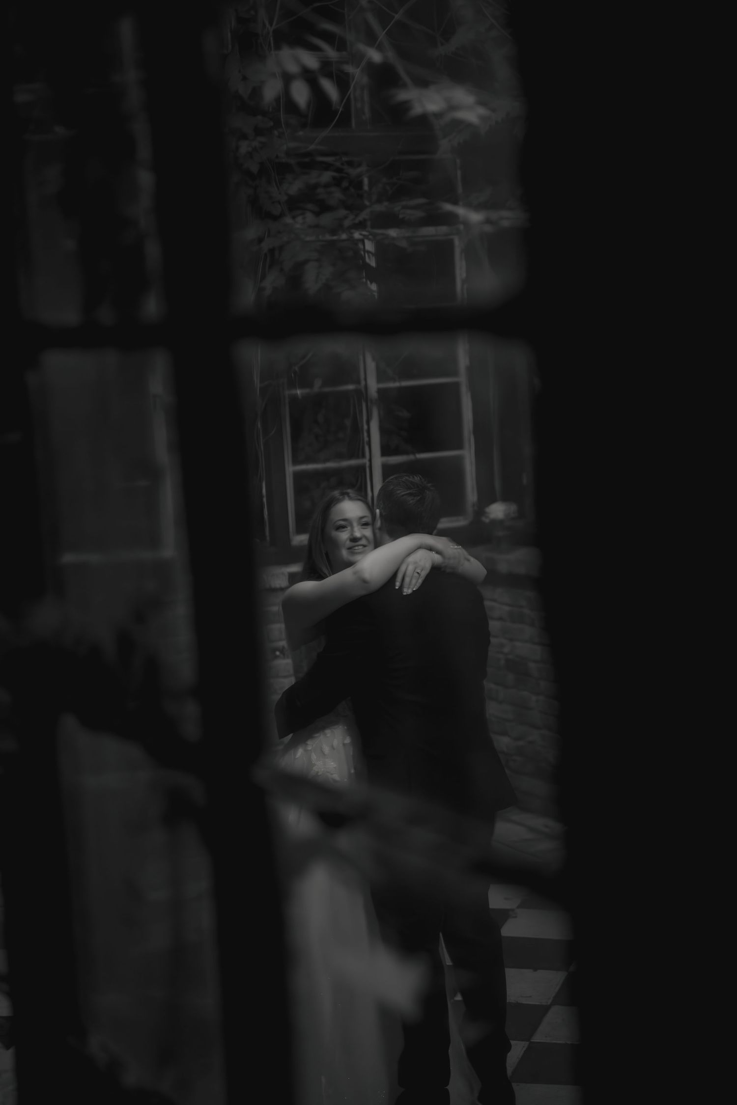 Intimate black and white photo of a couple embracing, viewed through a window frame.