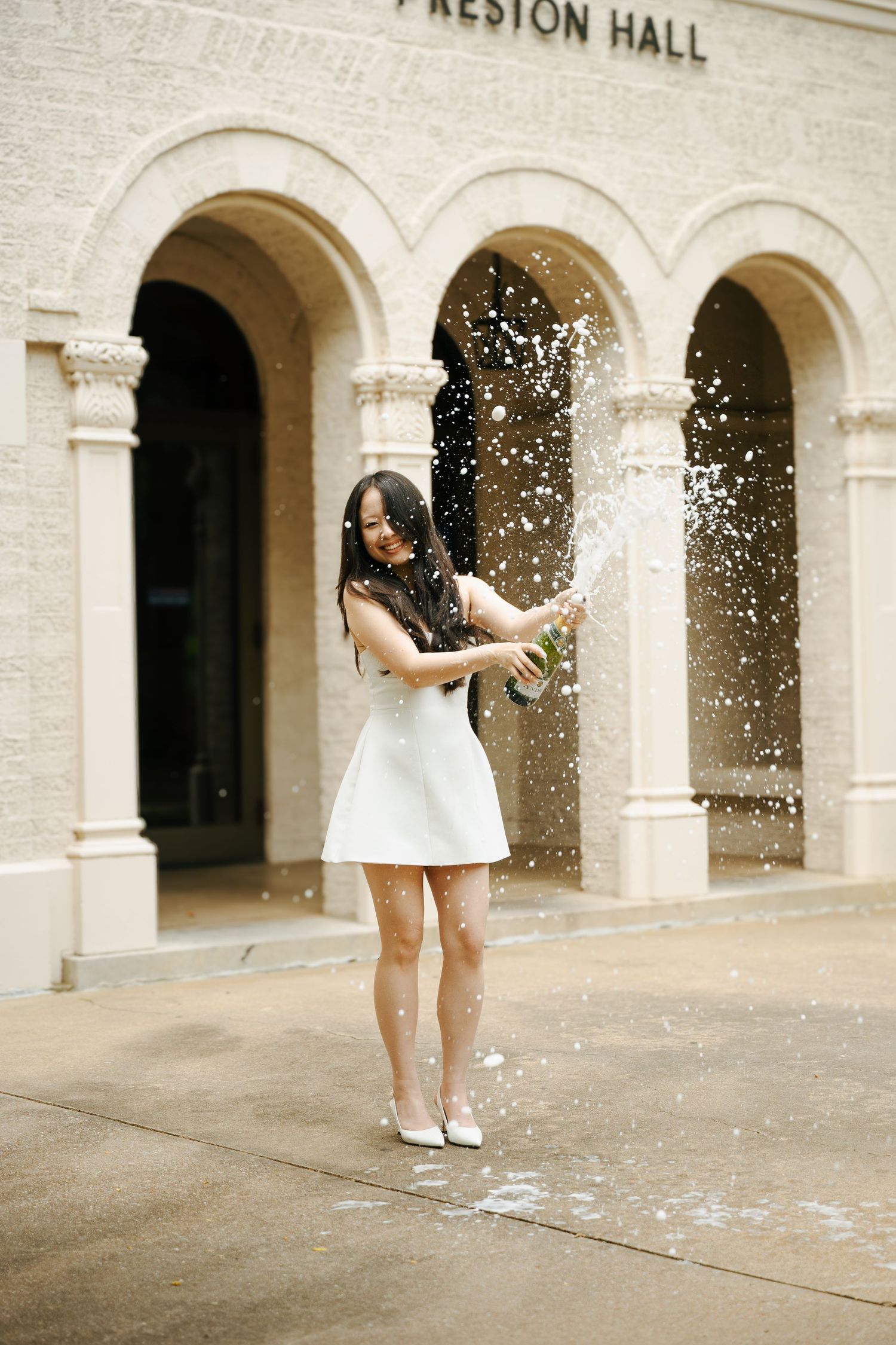 Woman in white dress popping champagne bottle outside Preston Hall building with arched stone facade.