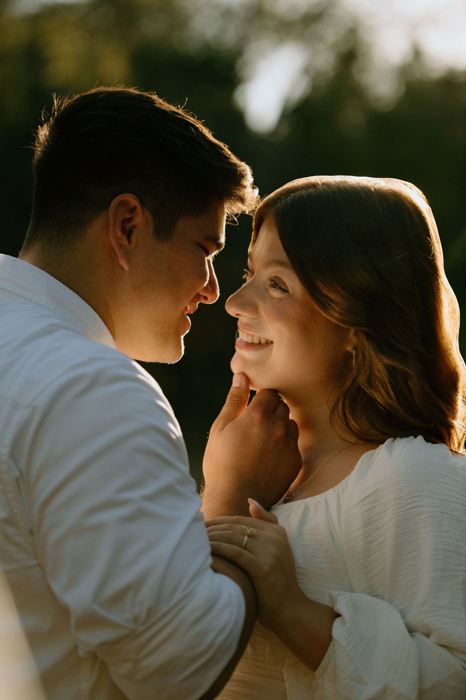 A romantic couple in white clothing share an intimate moment backlit by golden sunlight during sunset.