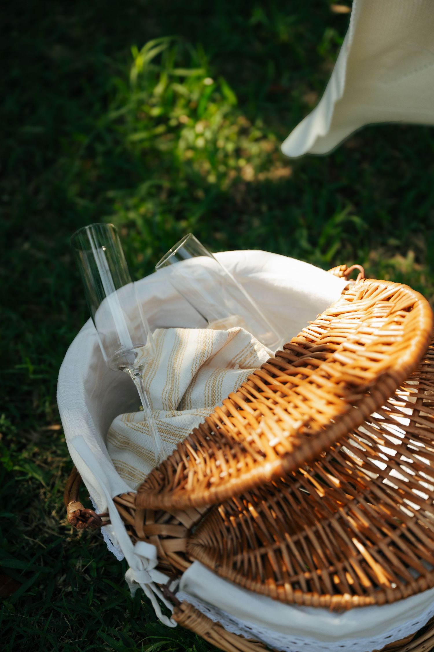 Open wicker picnic basket lined with white fabric, containing striped napkins and glassware, resting on green grass.