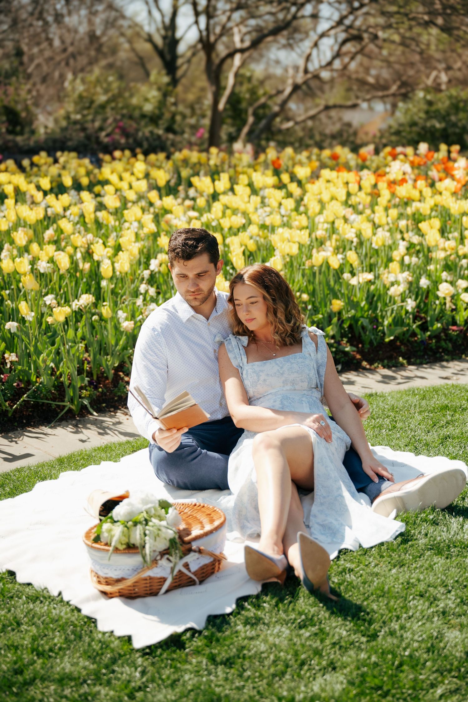 Couple enjoying a romantic spring picnic on a white blanket in front of vibrant tulip garden with a wicker basket.