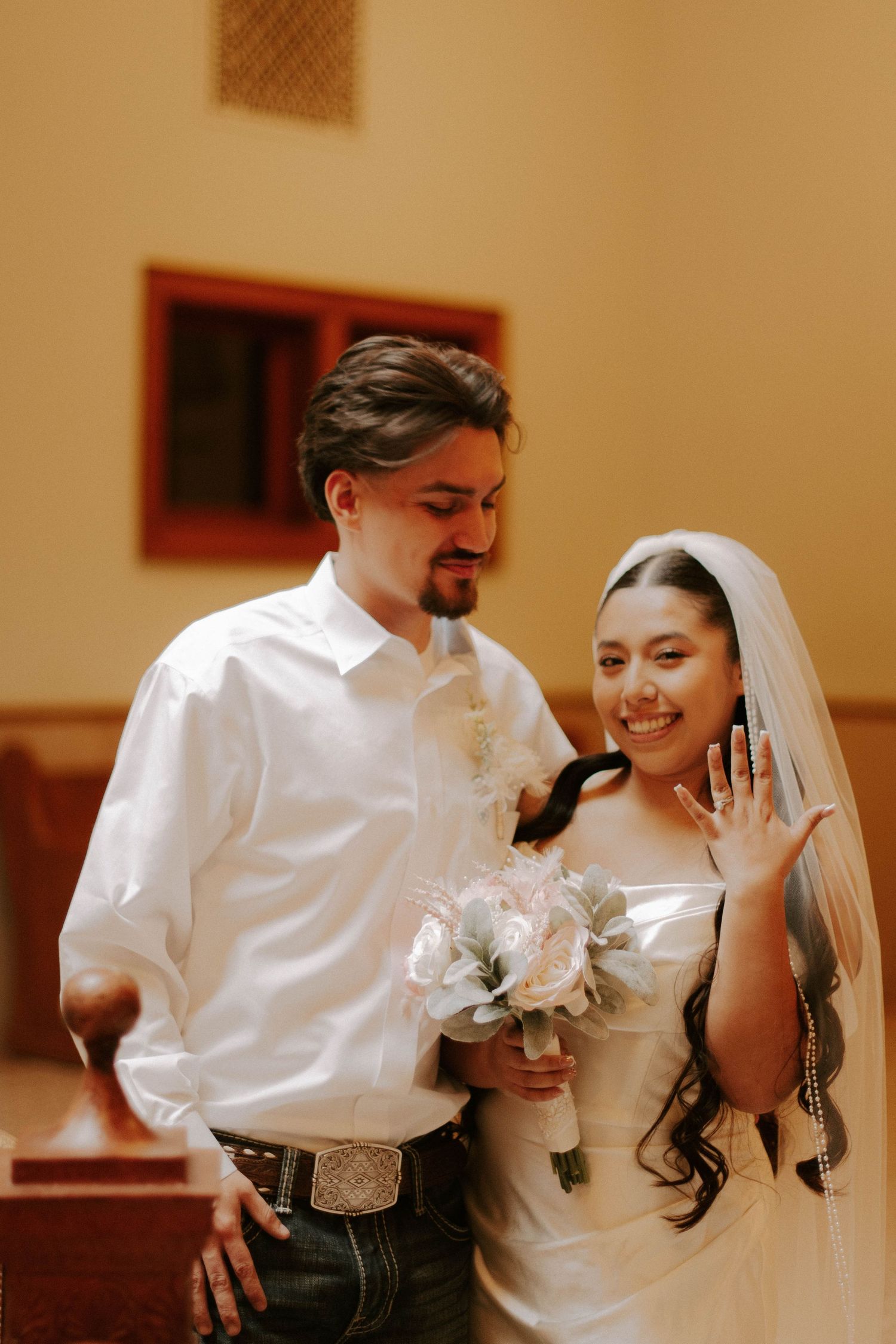 Newlywed couple smiling in formal attire with bride showing off her ring.