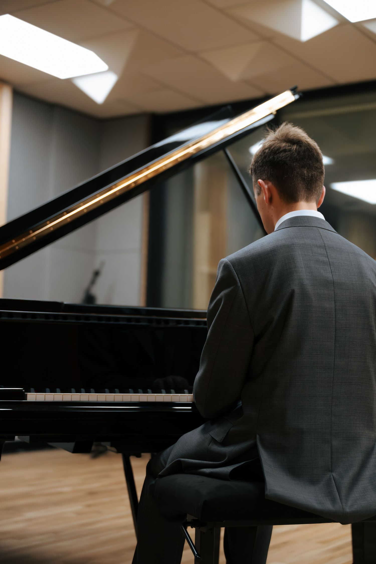 A man in a gray suit plays a black grand piano from behind in a professional studio setting.