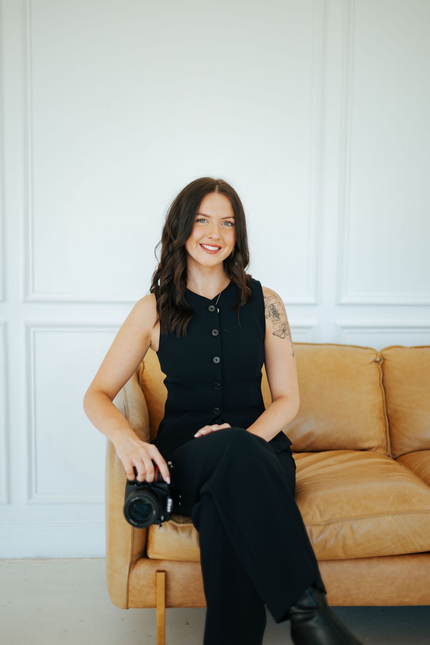 A person in a black sleeveless dress sits casually on a tan leather couch against a white wall.
