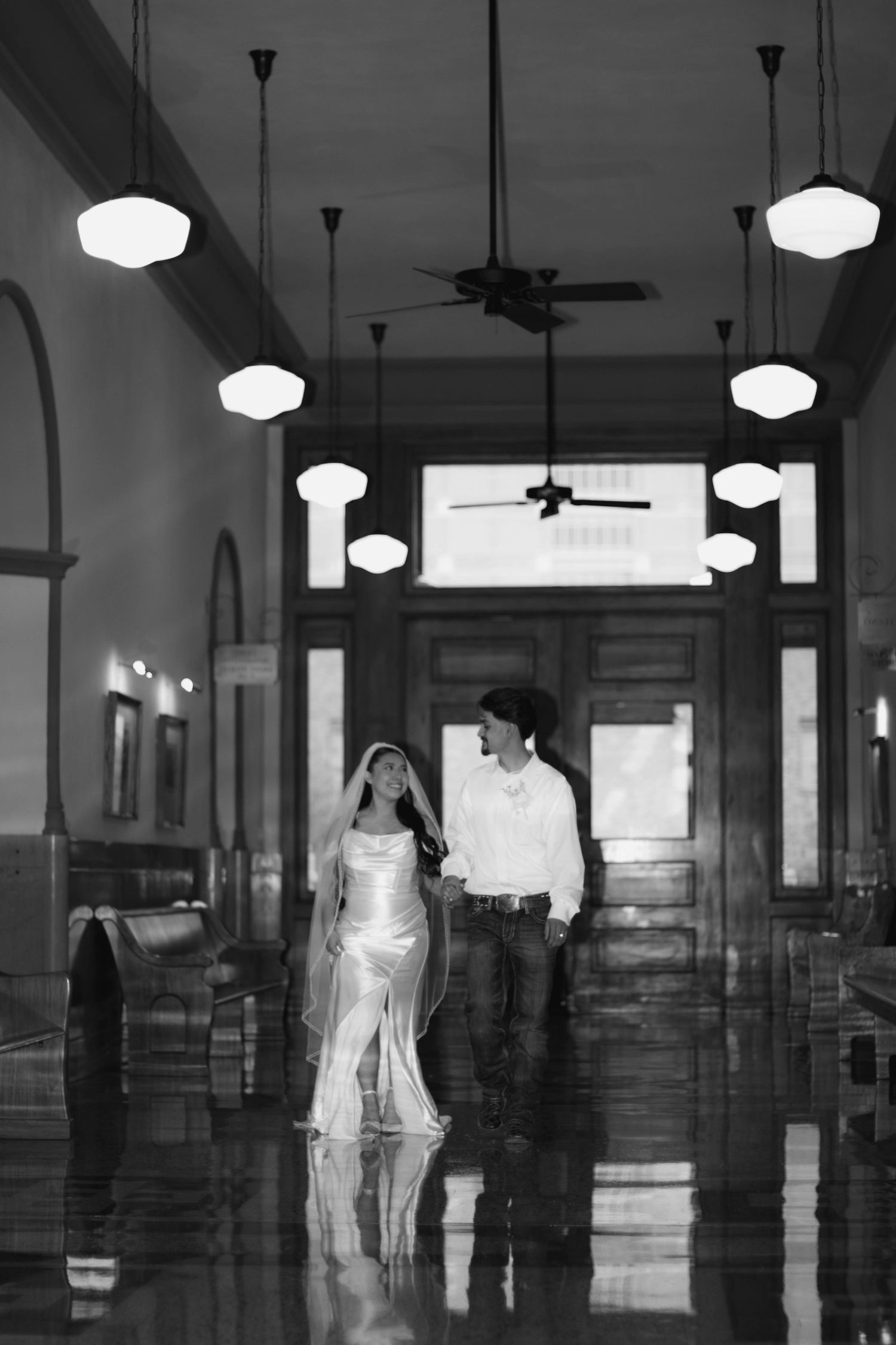 A couple walks through an elegant hallway with hanging pendant lights reflecting on the polished floor.