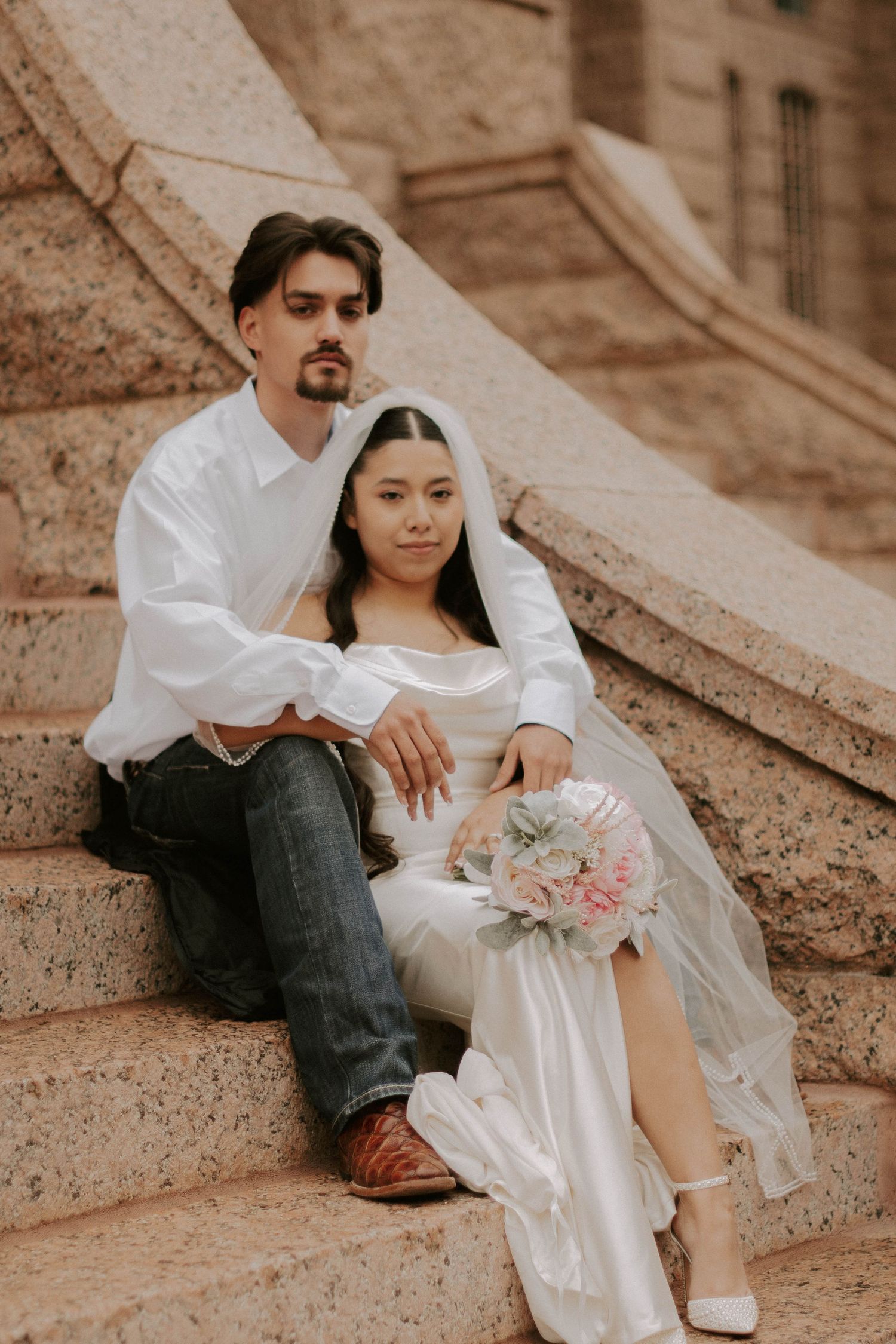 A couple in wedding attire sits on stone steps holding a delicate pink and white bouquet.
