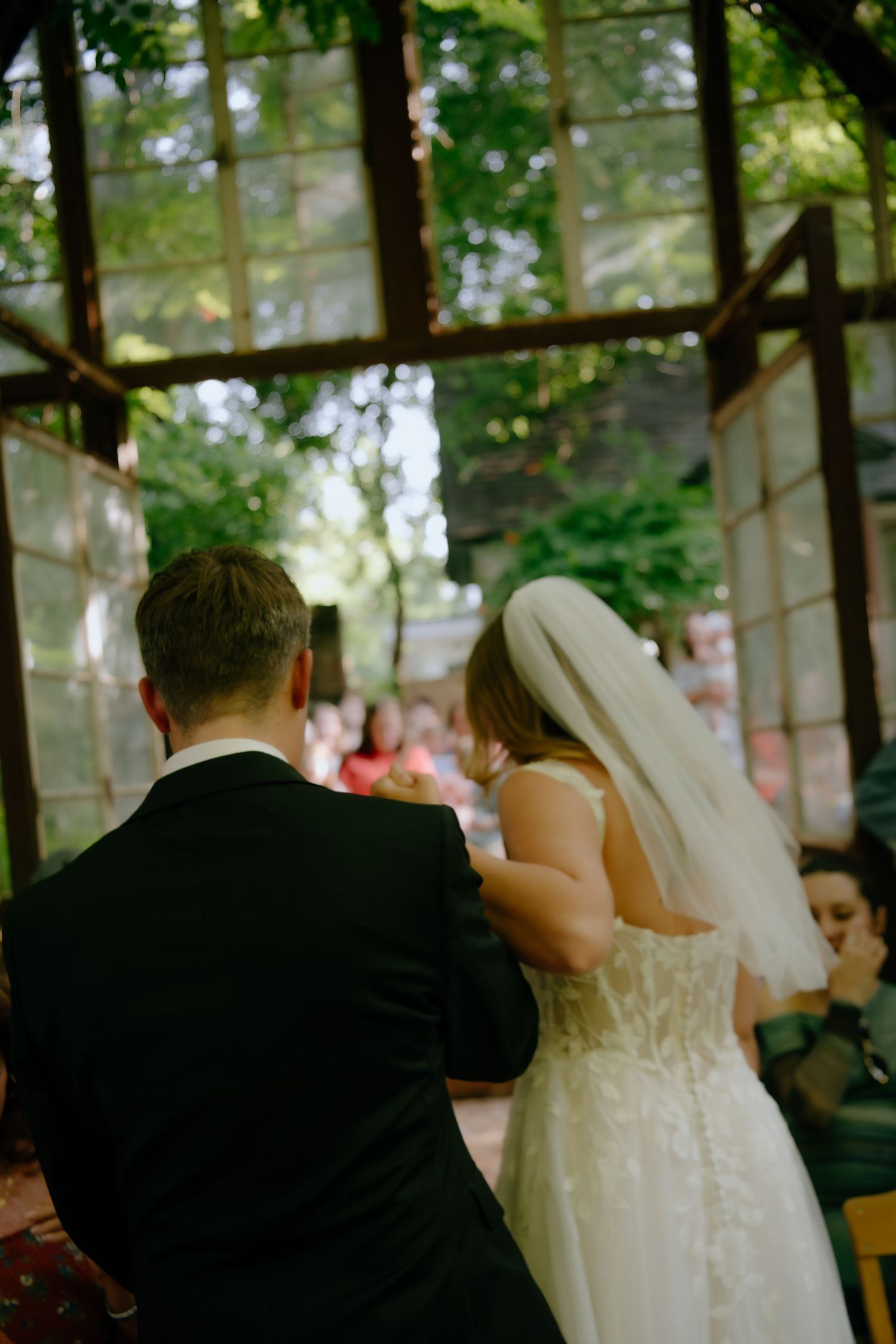 Wedding couple seated in a greenhouse with sunlight streaming through glass panels.